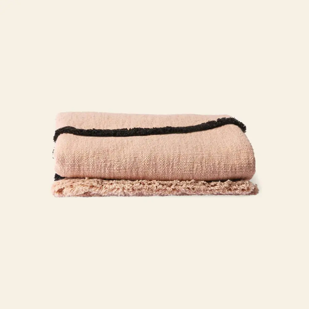 The HKliving Soft Woven Throw With Black Tufted Lines is a cosy cotton blanket in pink, featuring black stripes near the edge and a stylish texture, folded neatly on a plain, light-colored background.