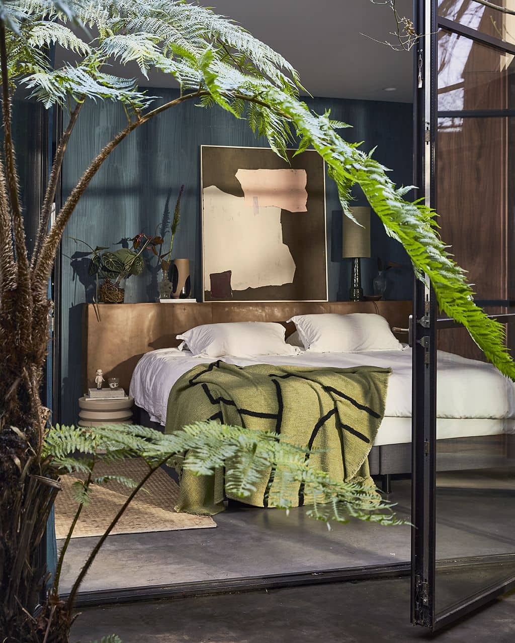 A stylish bedroom with a double bed, cosy white cotton bedding, and the HKliving Soft Woven Throw With Black Tufted Lines. Large plants frame the space, while abstract art and textured dark blue walls complete this naturally lit retreat.