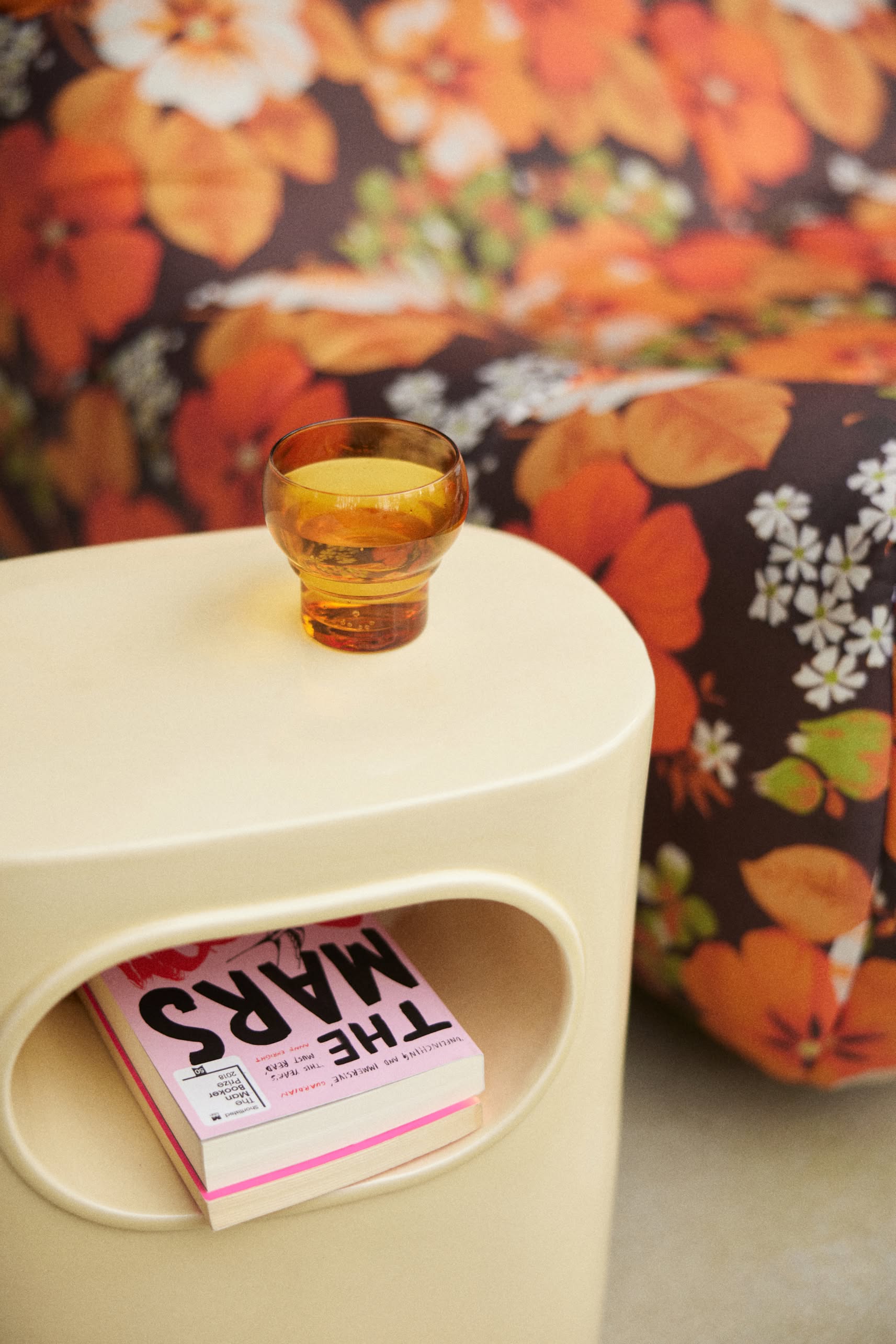 The HKliving Space Side Table by HKliving, in beige with a round opening, holds a pink THE MARS book and an amber glass cup. Behind it, a couch with a vibrant orange floral pattern brings modern flair to the room.
