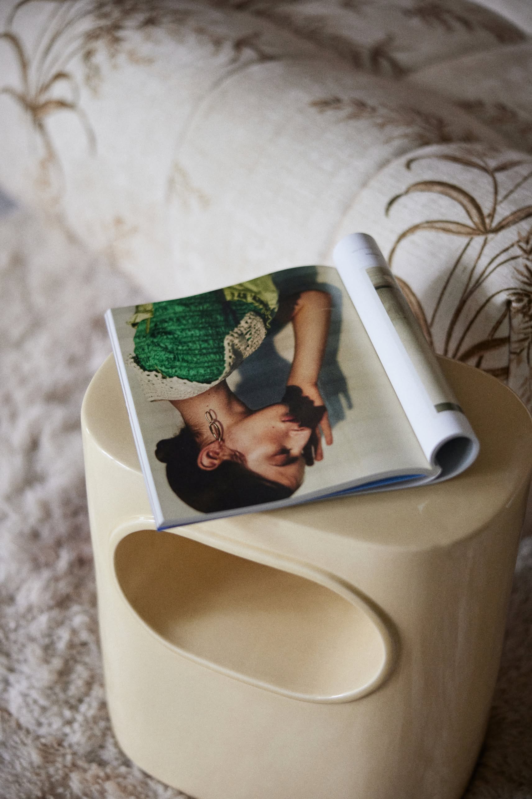 A magazine open to a page with a woman in a green top rests on the HKliving Space Side Table by HKliving, featuring a modern beige design and placed in front of a patterned couch.