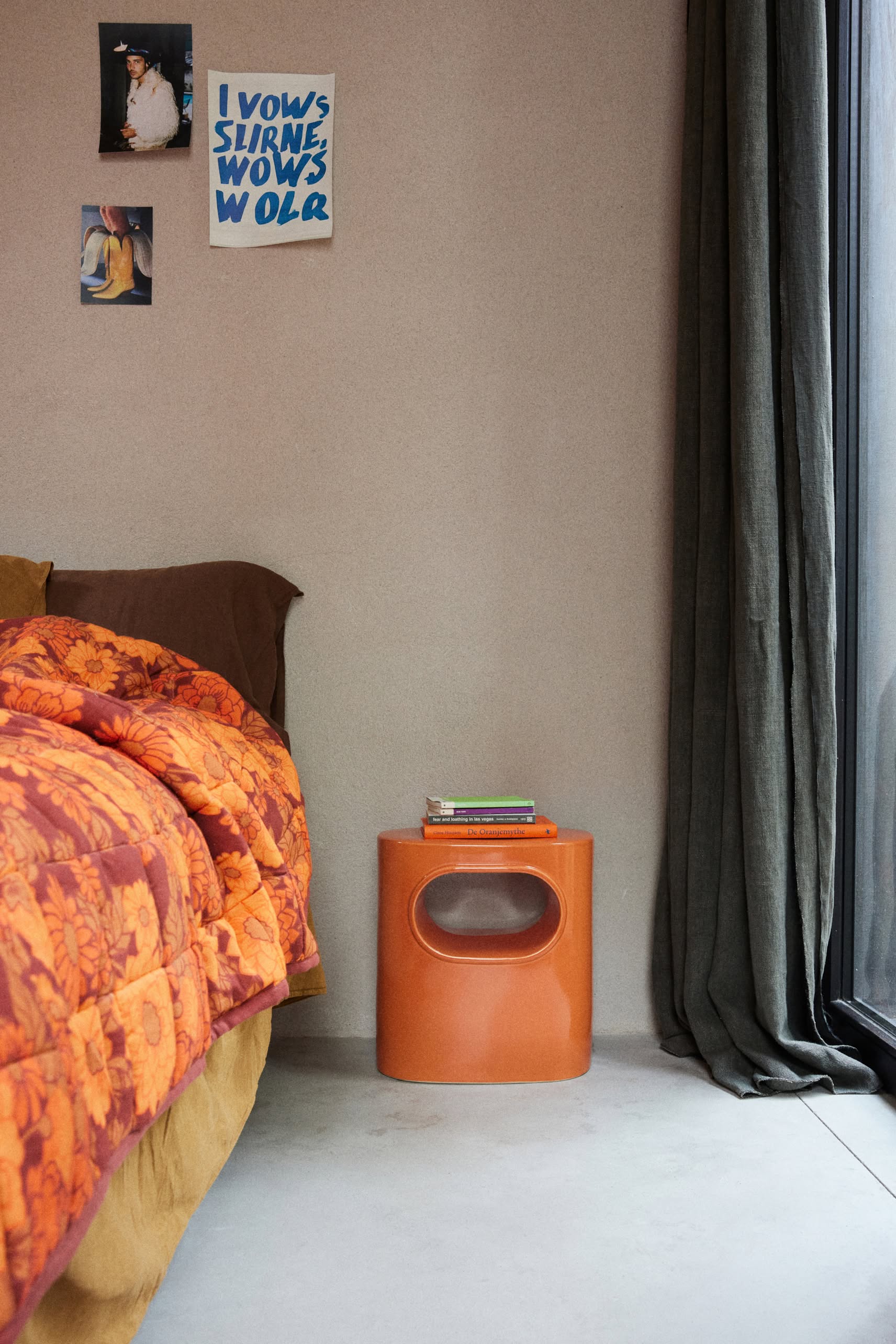 A cozy bedroom corner featuring the HKliving Space Side Table in orange retro style holding books, an orange floral bedspread, and three art prints above the bed, beside a large window with dark curtains.