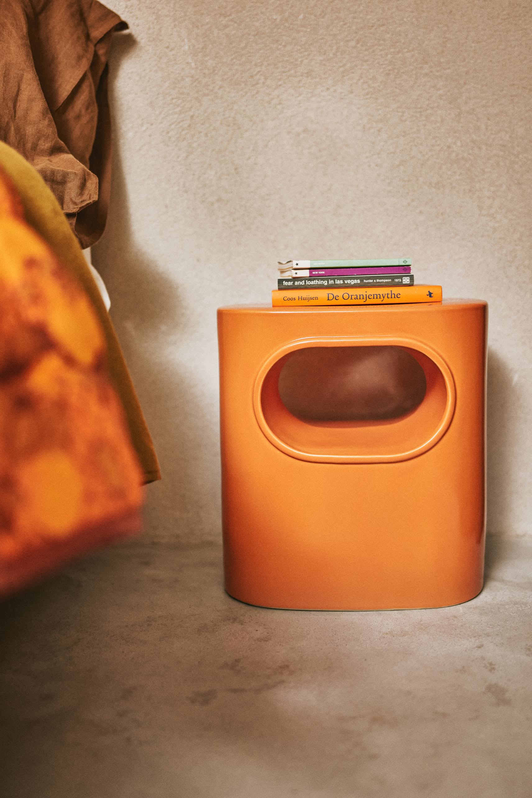 The HKliving Space Side Table by HKliving, with an orange oval design and built-in handle, sits on a light carpet topped with three stacked books. An orange and yellow blanket appears in the left foreground.