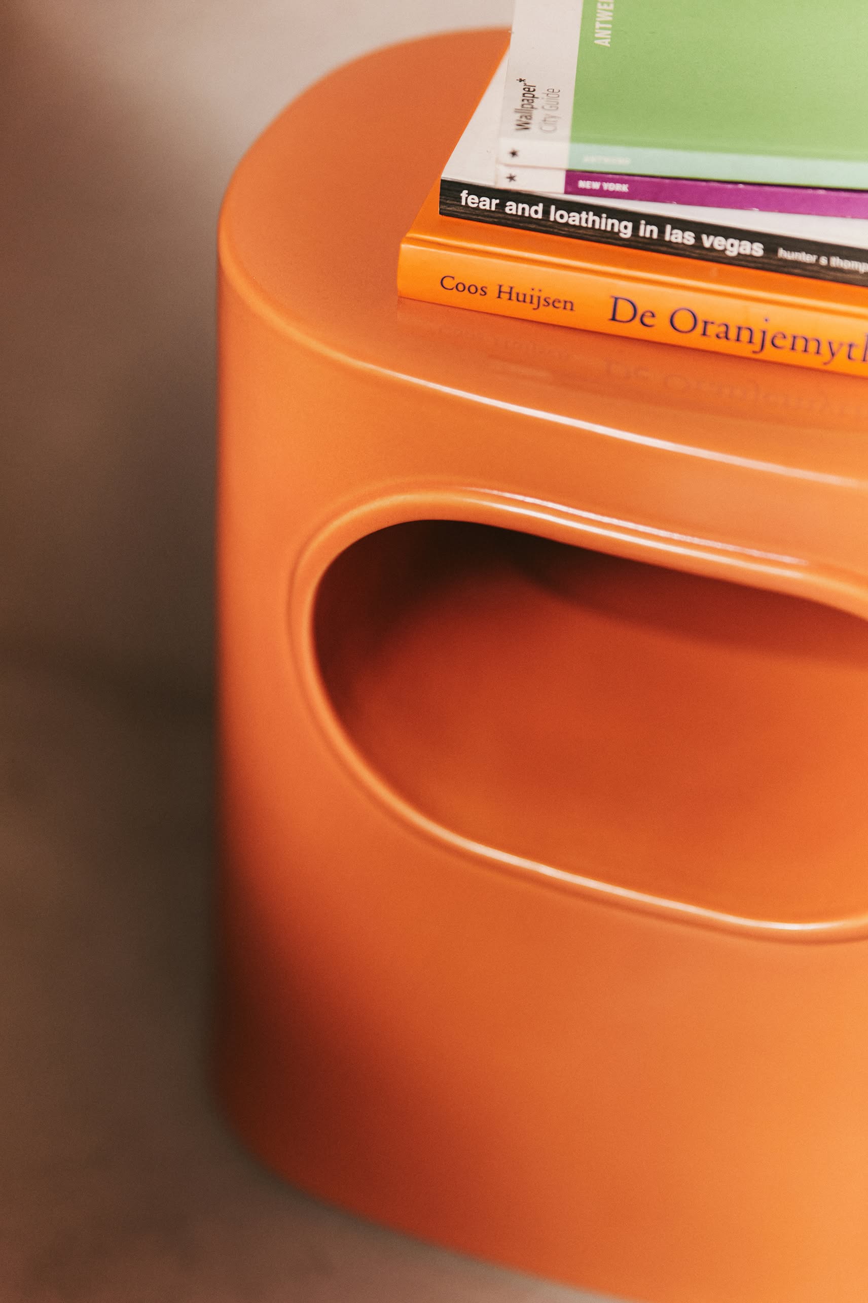 A close-up of the HKliving Space Side Table by HKliving showcases its orange color, oval cutout, modern design, and stacked books—De Oranjemythe and Fear and Loathing in Las Vegas—emphasizing its multifunctional storage.