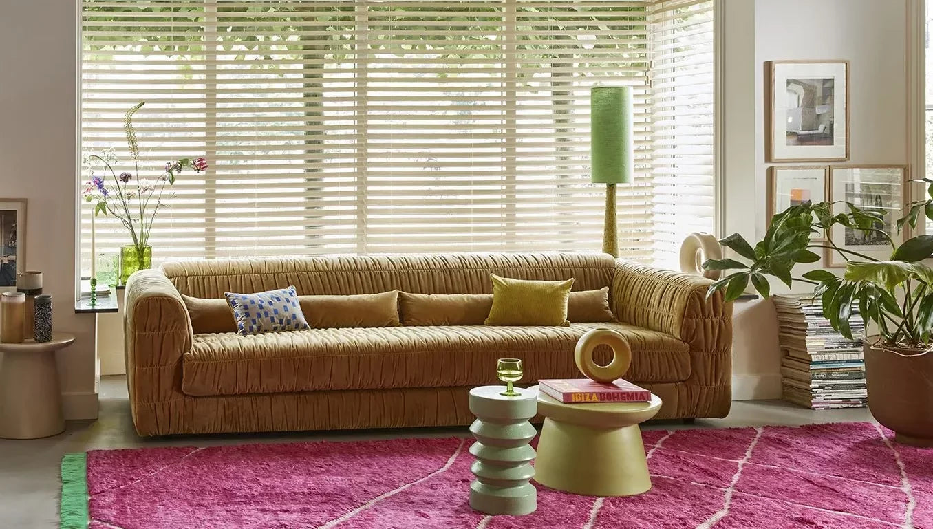 A cozy living room with a plush tan sofa, colorful pillows, a magenta rug with white lines, two modern coffee tables including the HKliving Stoneware Side Table by HKliving for whimsical charm, houseplants, books, and sunlit windows with blinds.