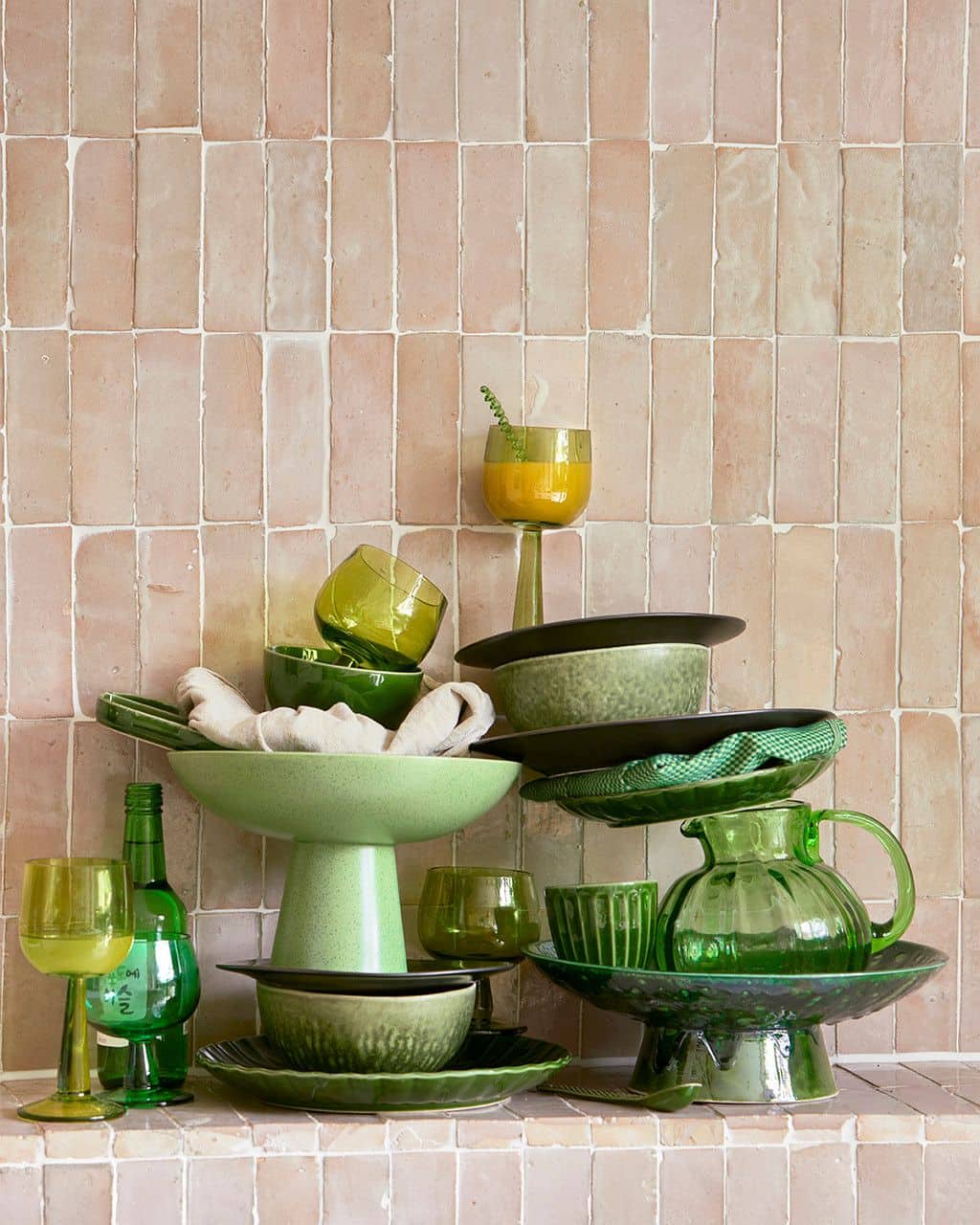 A stack of green glassware and dishes, including the HKliving The Emeralds: Ceramic Mug Ribbed (Set of 4), is arranged against pale pink tiles with a beige cloth draped in one bowl.