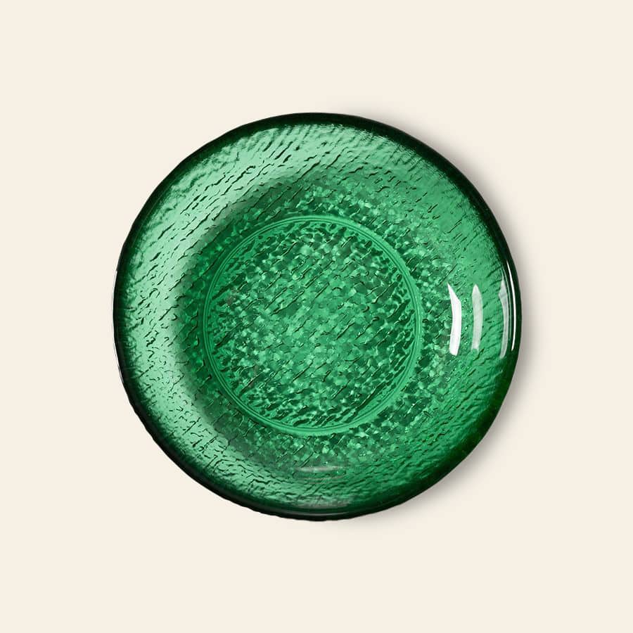 A round green glass plate with a textured pattern is shown from above on a light beige background, reflecting the modern elegance of HKliving’s The Emeralds: Glass Dessert Bowl collection.