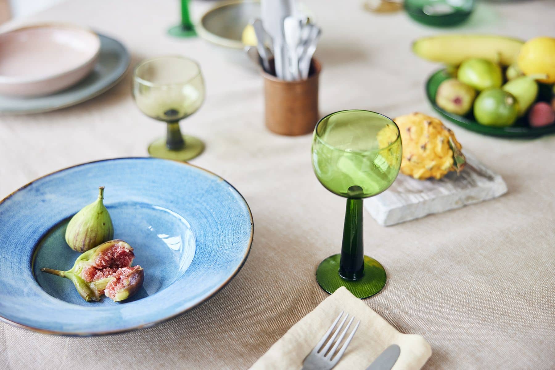 HKliving’s The Emeralds: Glass Dessert Bowl in green adds modern elegance to a table set with a blue plate of figs, utensils on a napkin, and assorted fruits like bananas and grapes atop a beige cloth.