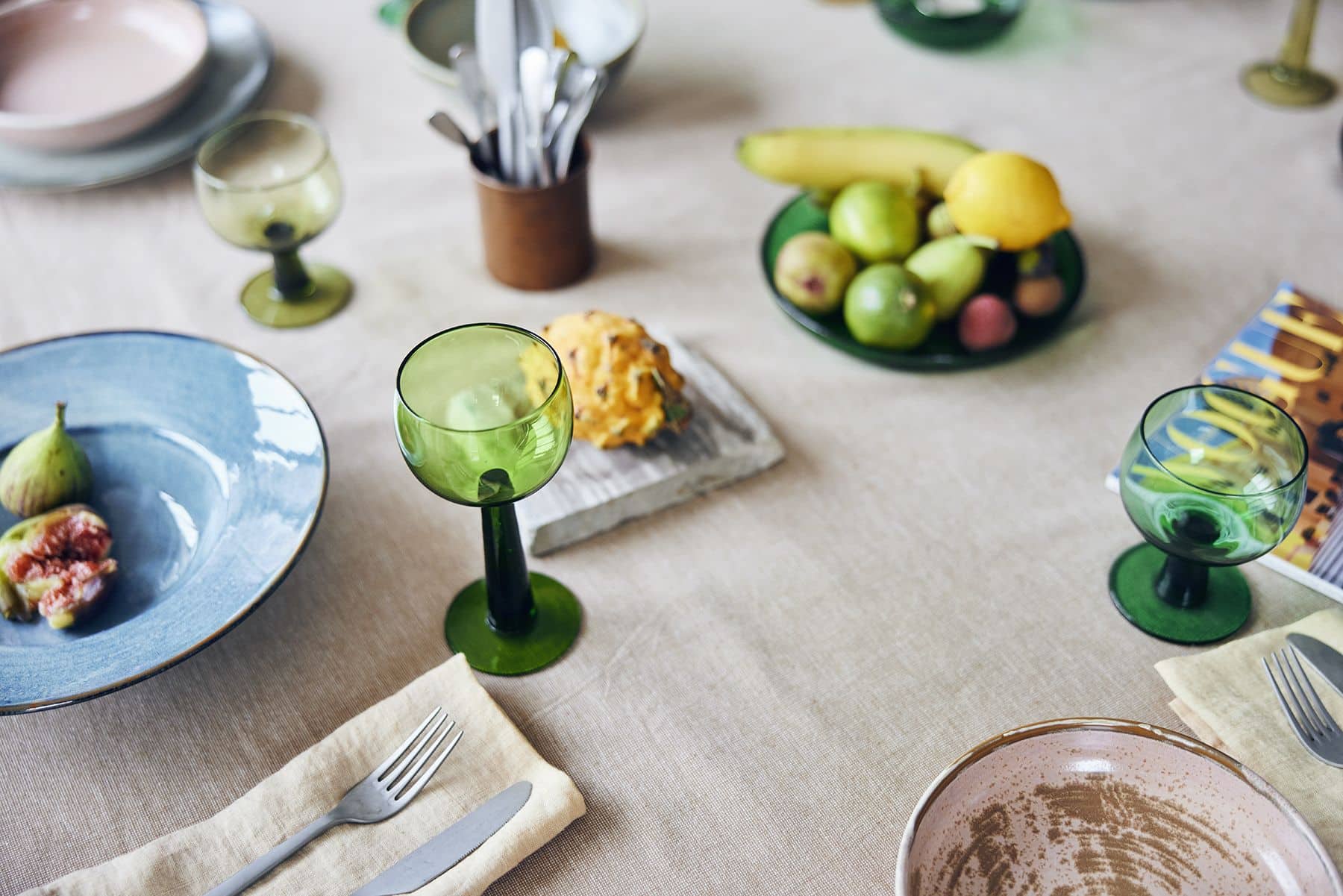 A dinner table set with HKliving The Emeralds: Glass Side Plates, green glass goblets, plates with figs, cutlery on napkins, and a fruit bowl of bananas, apples, and a lemon on a beige tablecloth.