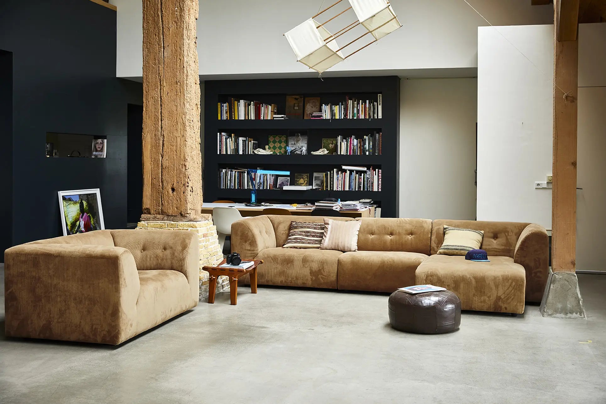 A modern living room features an HKliving Vint couch with customizable seating, an HKliving hocker, a wood side table, exposed wooden beams, and a black bookshelf filled with books against a dark wall.