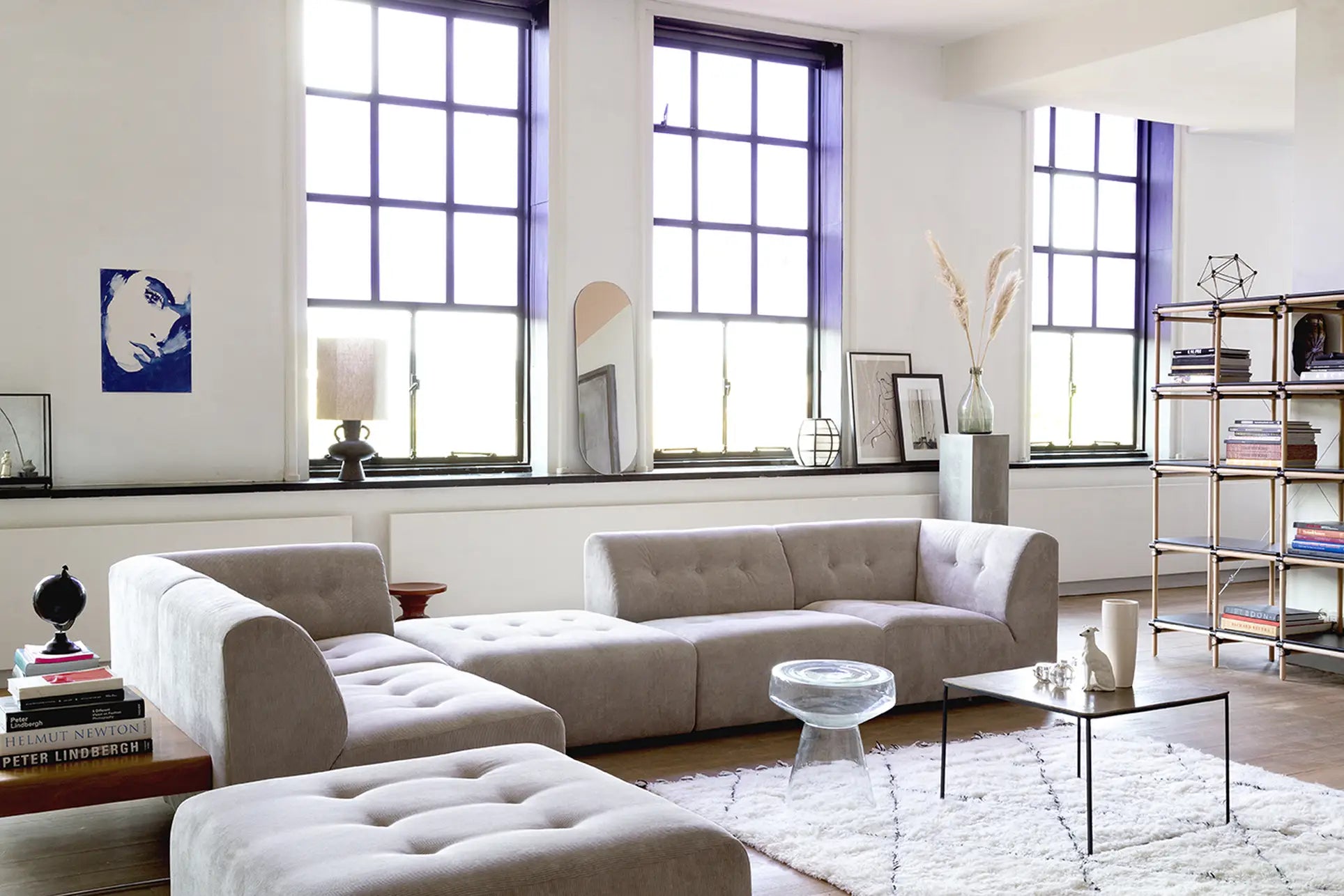 A modern living room with large windows, a light gray HKliving Vint Couch and Hocker featuring customizable seating, a glass side table, black coffee table, white rug, wall art, and tall shelving. The airy space is styled with minimalist decor.