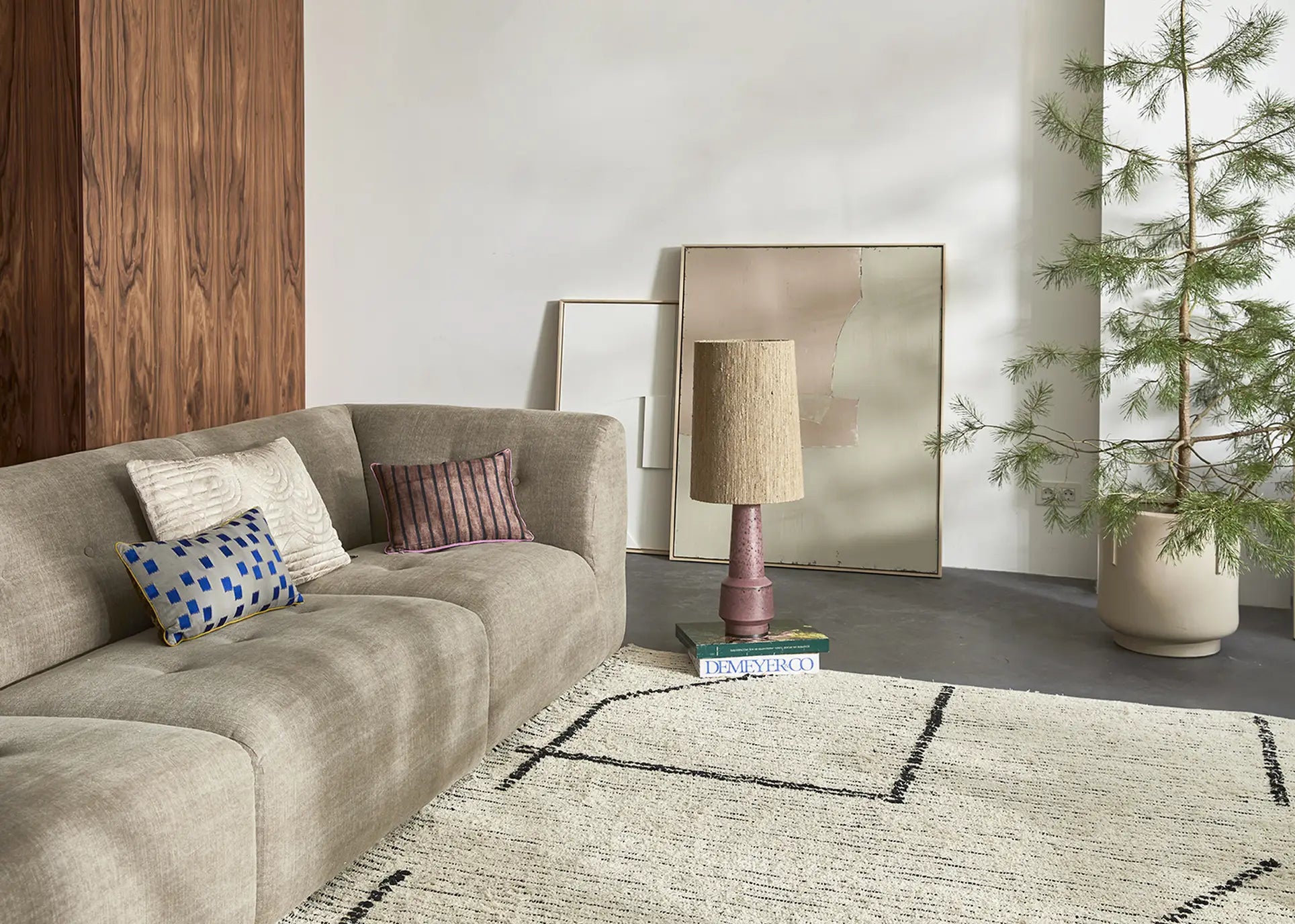 A modern living room showcases the HKliving Vint Couch and Hocker in beige with colorful cushions, a textured rug, abstract art on wood paneling, a potted tree, and a floor lamp resting on stacked books.
