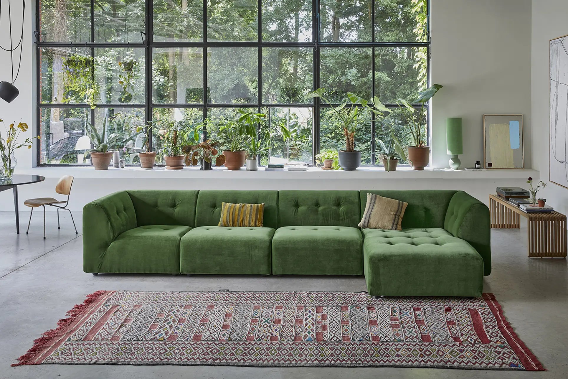 A modern living room with the HKliving Vint Couch and Hocker Small, a patterned red rug, potted plants on a white windowsill, and floor-to-ceiling windows displaying lush greenery for a customizable space.