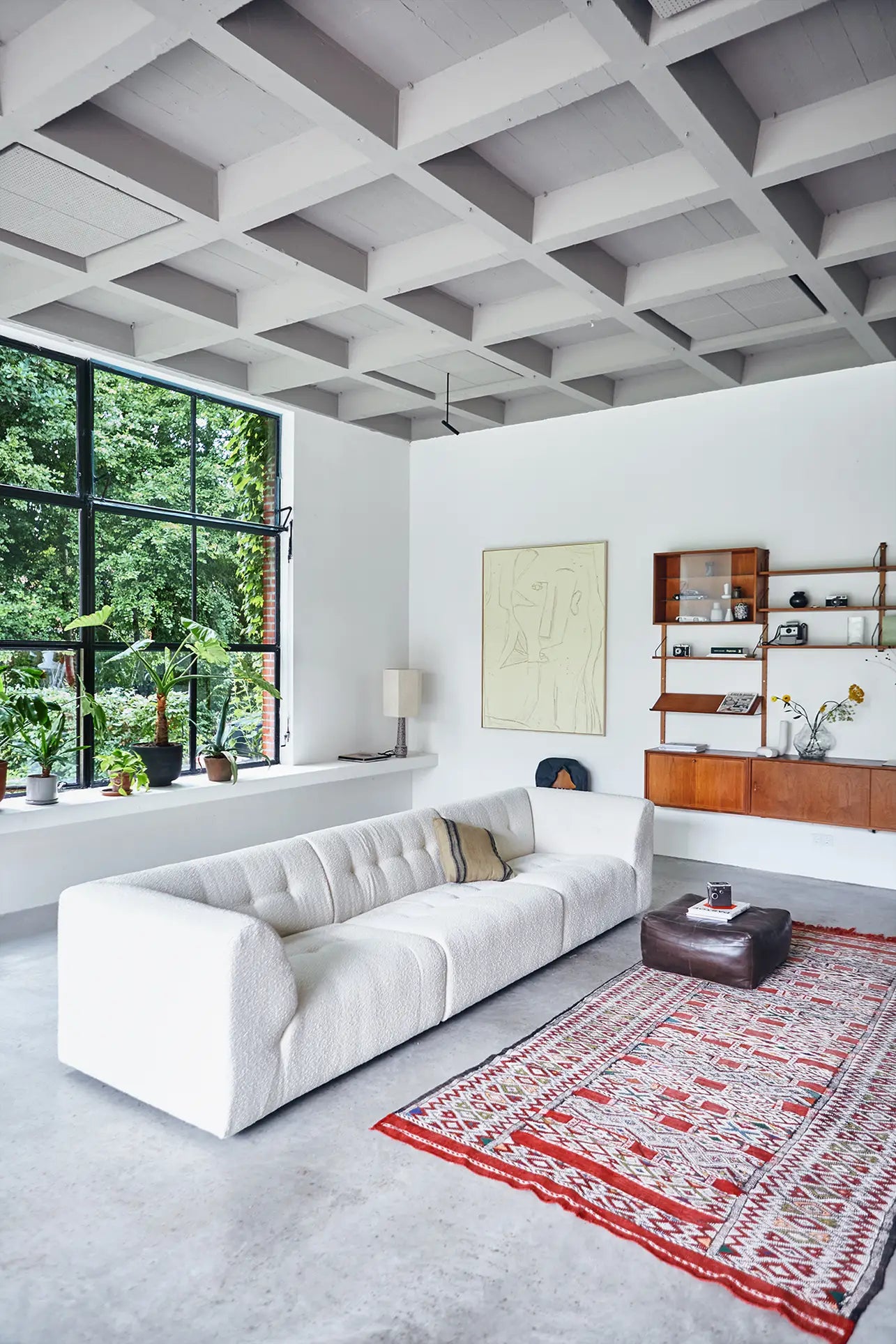 A modern living room featuring the HKliving Vint Couch, Left, 1.5-Seat in white, a red patterned rug, brown ottoman, wall-mounted wooden shelf, black-framed windows, indoor plants, and a gray coffered ceiling creates a customizable space.