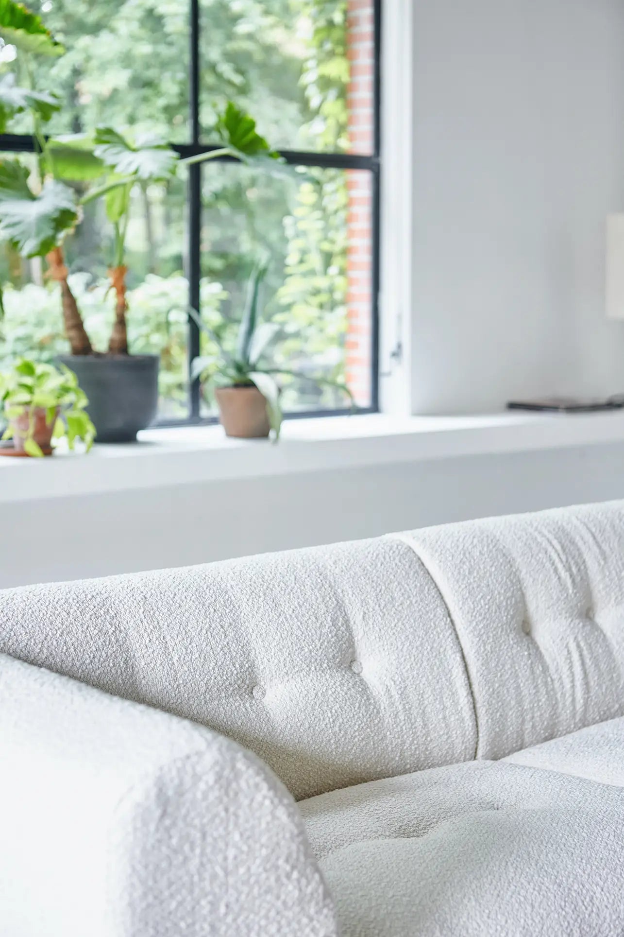 A close-up of the HKliving Vint Couch, Left, 1.5-Seat in white textured fabric by HKliving sits by a windowsill with green potted plants and natural light pouring in—an ideal piece to personalize your living space.