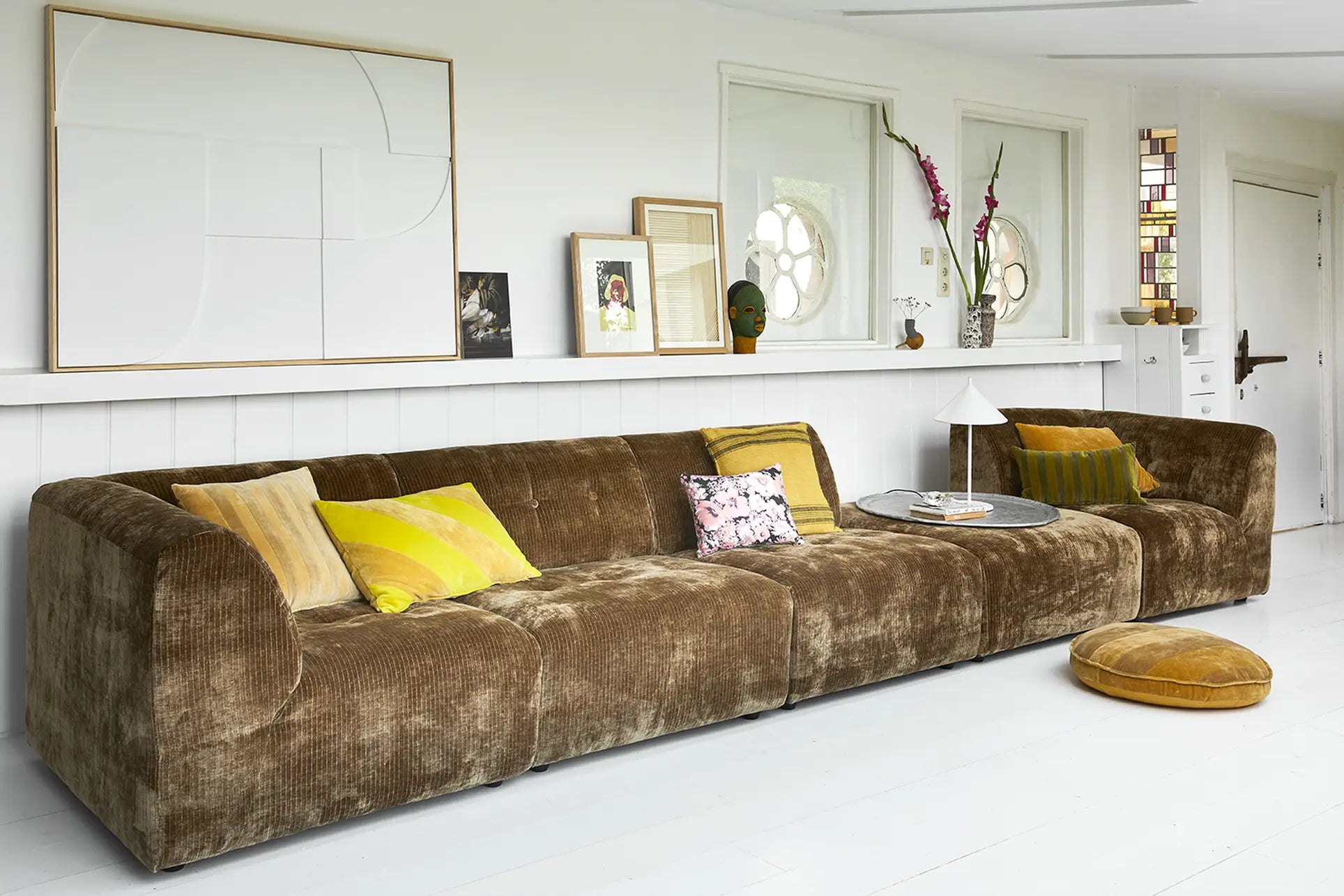 The HKliving Vint Couch, Left, 1.5-Seat by HKliving features a long brown design with colorful cushions, set in a bright modern living room with white walls, art, decor pieces, and a round yellow floor cushion for customizable style.