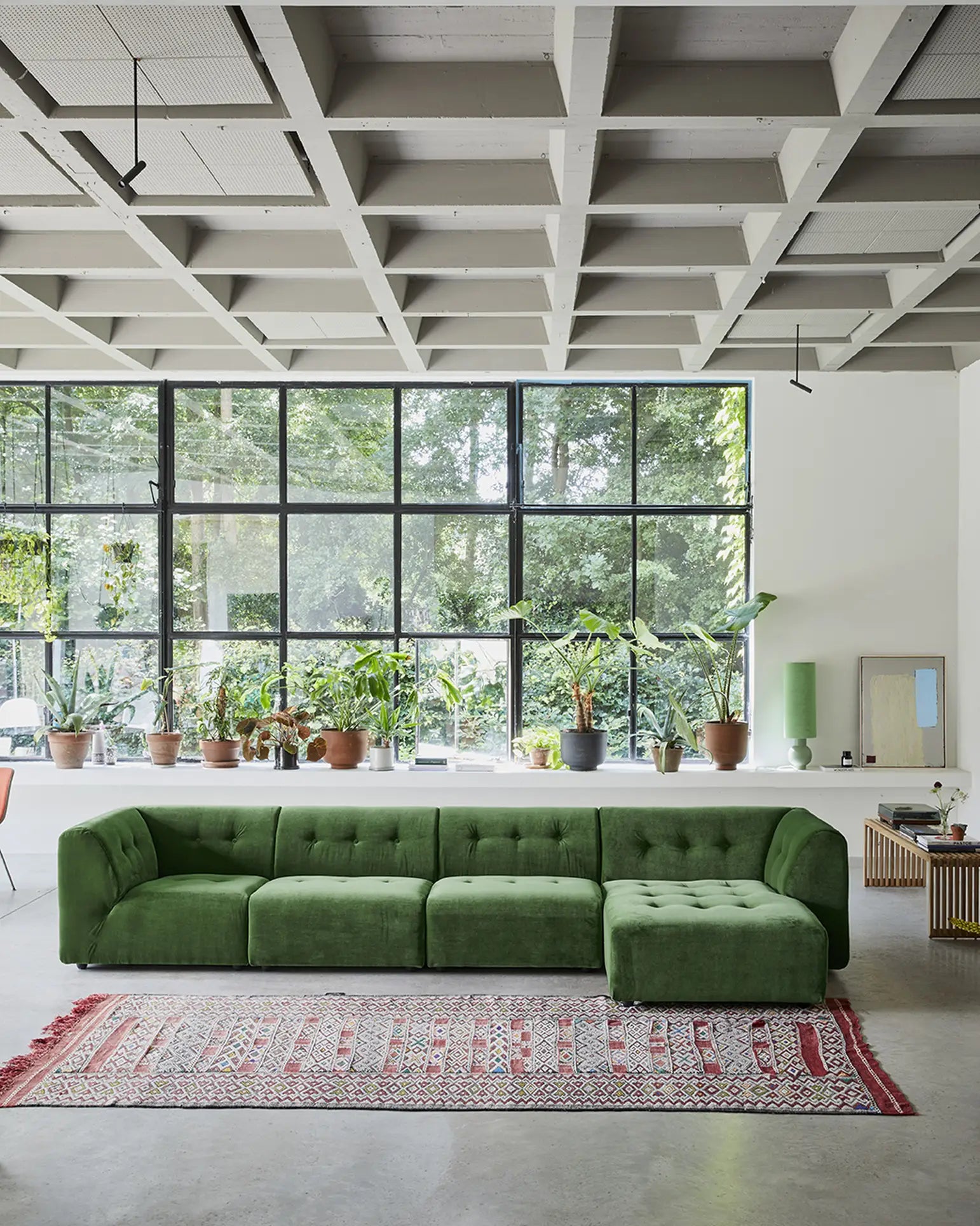 A modern living room with the HKliving Vint Couch, Left, 1.5-Seat—a spacious green sofa—adaptable components, a patterned red rug, and potted plants by tall windows. Exposed ceiling beams lend an industrial touch to this customizable space.