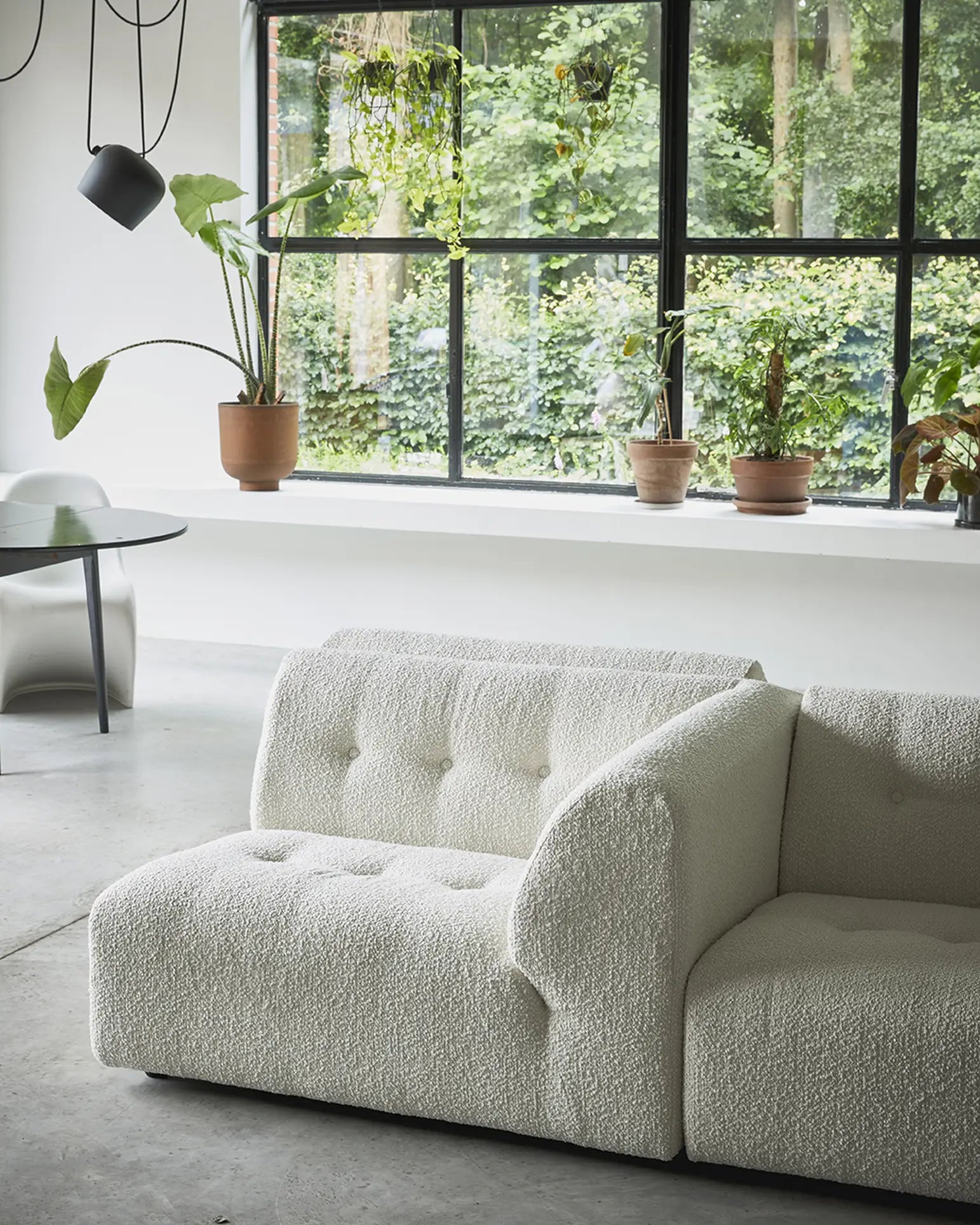 A modern living room features the HKliving Vint Couch, Left by HKliving, large black-framed windows with potted plants on the sill, and a round glass dining table with a gray chair in the background. Lush greenery is visible outside.