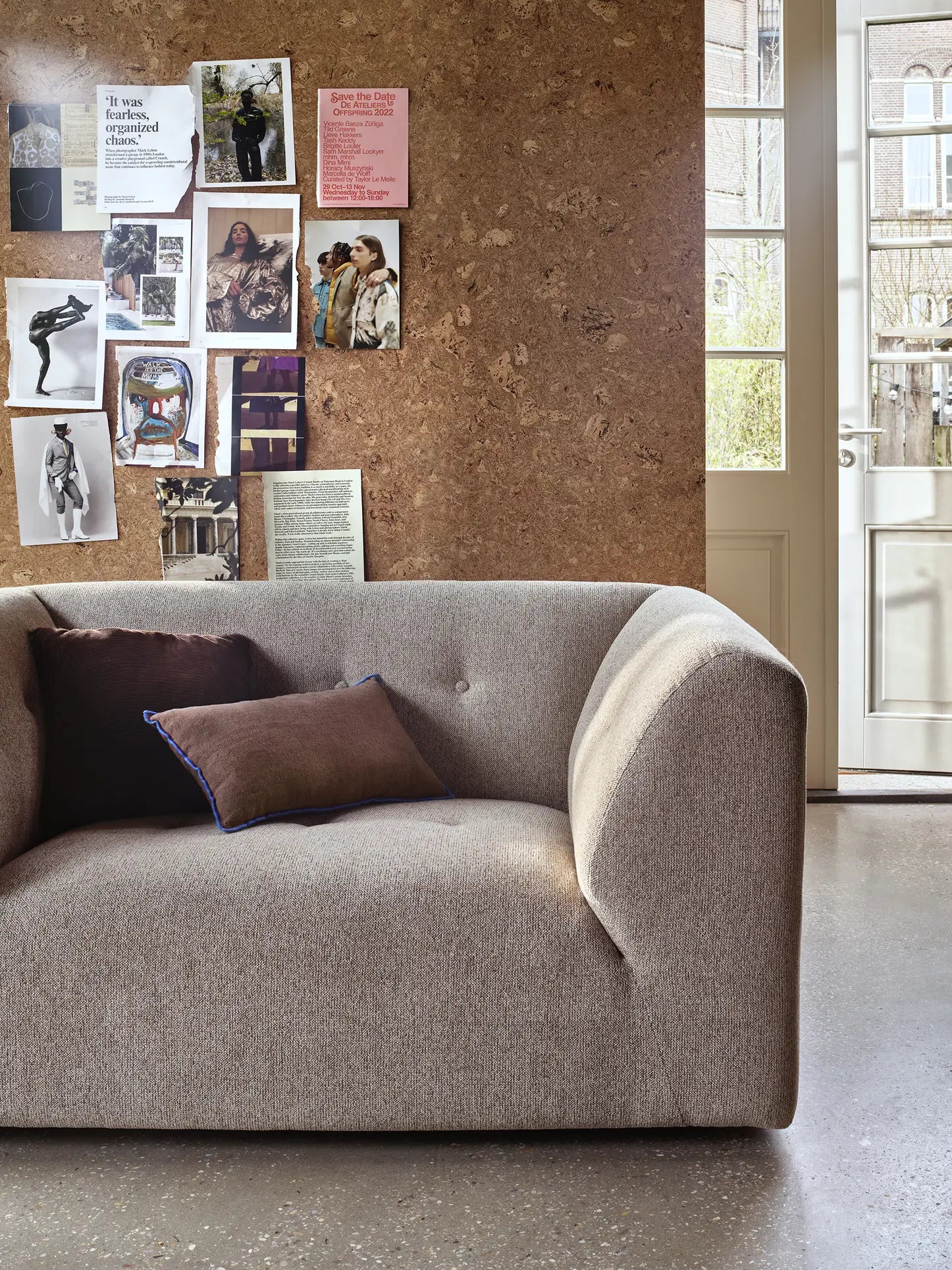 Modern living room with a light gray HKliving Vint Couch Loveseat, brown and purple pillows, corkboard wall with photos and notes, and a glass door that fills the customizable space with natural light.