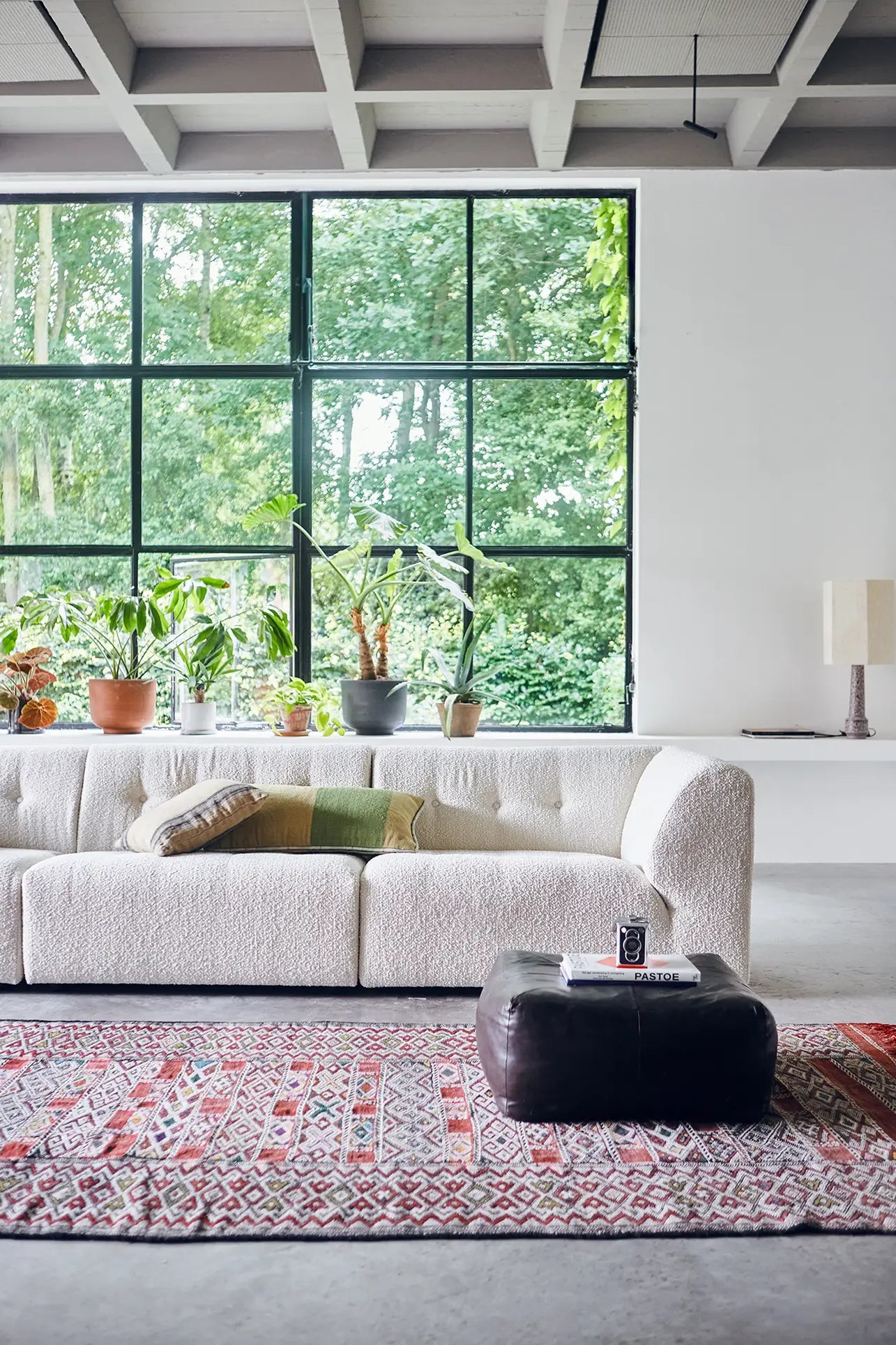 A modern living room with a large window, an HKliving Vint Couch, Middle with customizable seating and cushions, a patterned red rug, black ottoman, indoor plants on the windowsill, a lamp on a side table, and green trees visible outside.