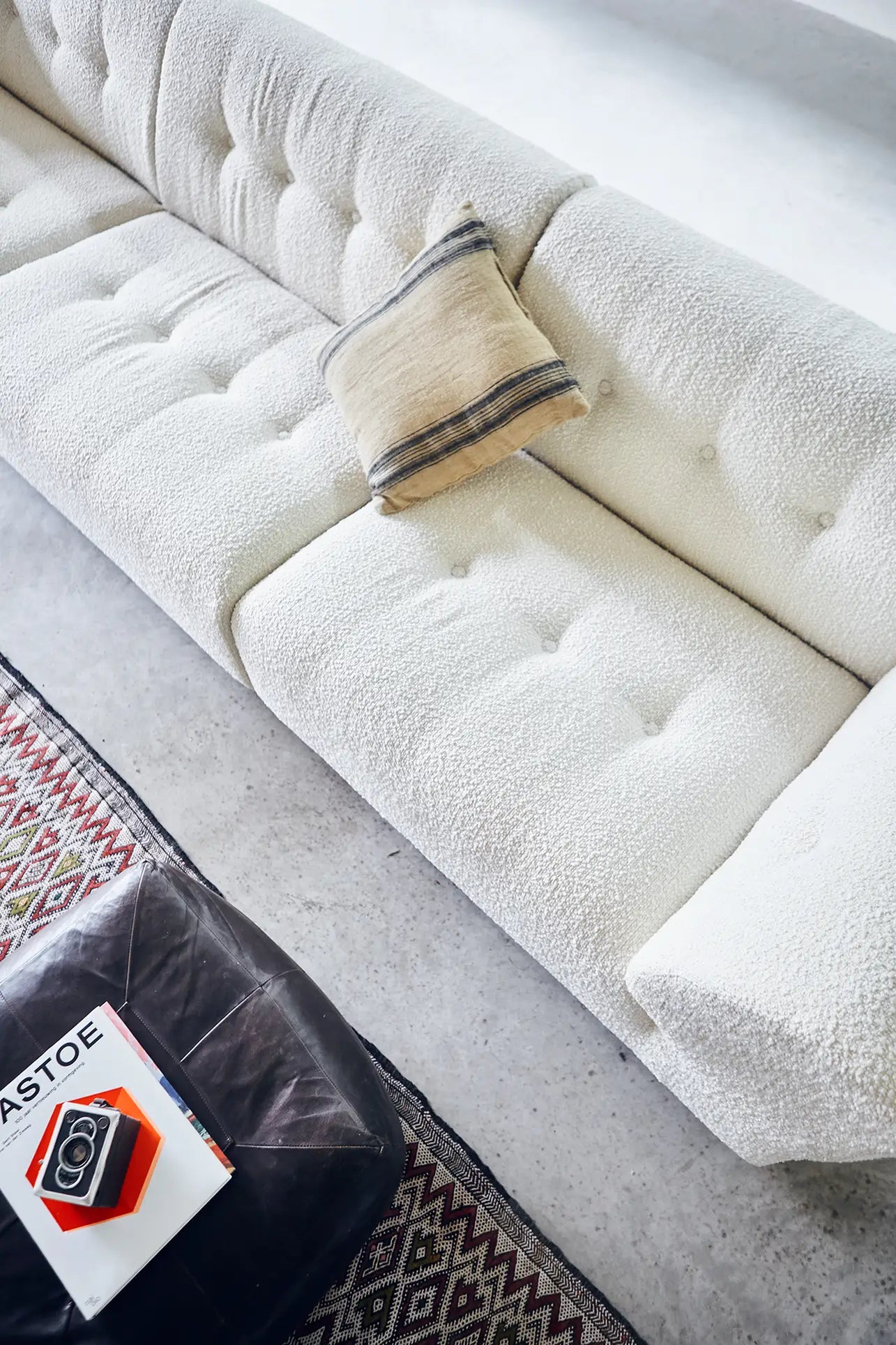 A top view of the HKliving Vint Couch, Middle in textured white fabric with a beige pillow, beside a patterned rug and dark coffee table with books and a red accent—ideal for showcasing HKliving’s customizable seating in any space.