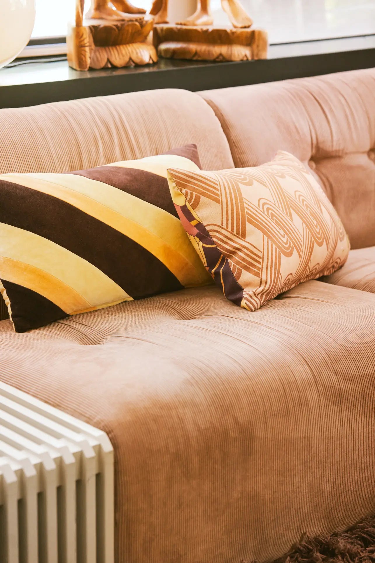 The HKliving Vint Couch, Middle, in light brown corduroy comes with two accent pillows—one striped, one with a wavy geometric pattern—creating customizable seating by a window featuring wooden decor.