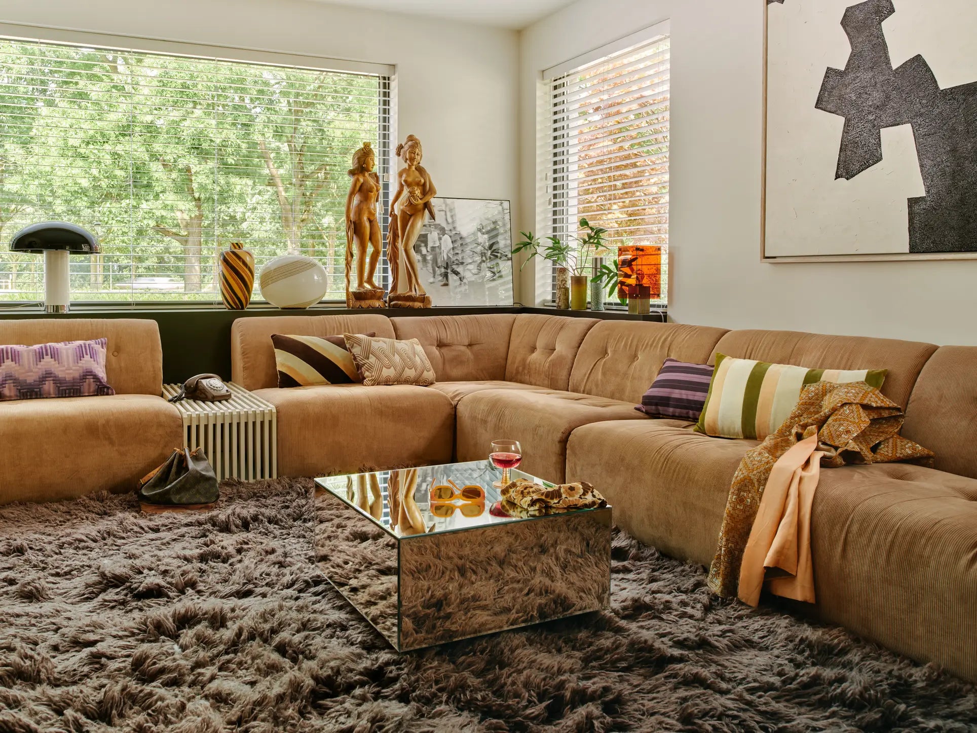 A cozy living room with an adaptable HKliving Vint Couch, Middle in tan, fluffy gray rug, mirrored coffee table with snacks and drink, large windows, contemporary decor, sculptures, and throw pillows in a bright, inviting space.