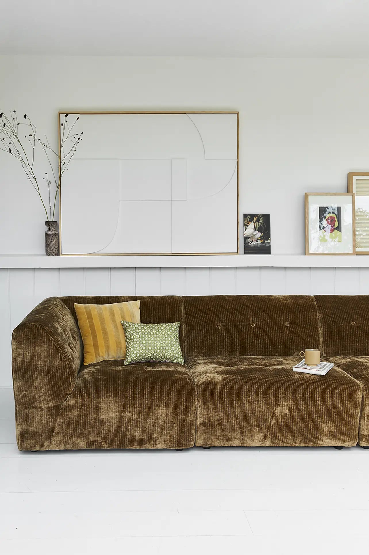 An HKliving Vint Couch, Middle in brown with customizable seating and two patterned pillows is placed in a bright living room. A shelf behind it holds framed artwork and a vase with dried branches; a cup with saucer sits on the sofa.