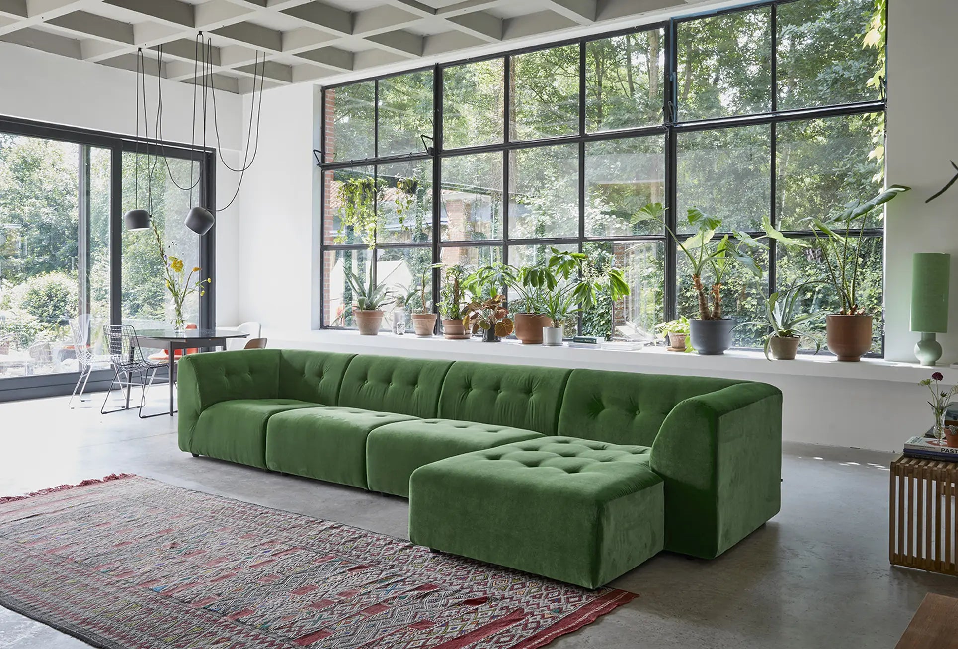A modern living room with the customizable HKliving Vint Couch, Middle by HKliving, a red patterned rug, and potted plants by windowed walls. Sunlight fills the space, highlighting garden views through the large windows.