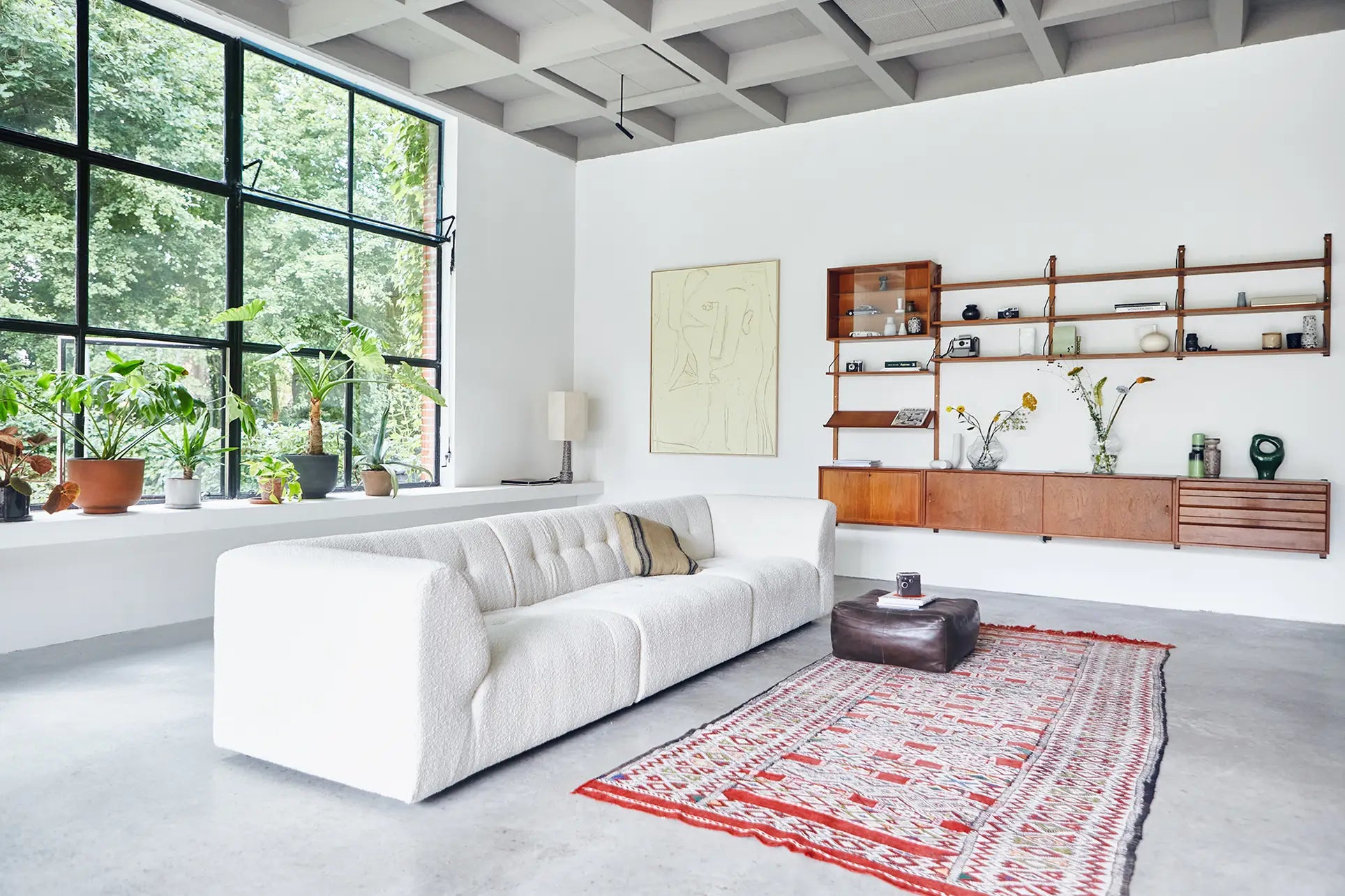 A bright living room with large windows, the HKliving Vint Couch, Right, a patterned red rug, potted plants, and a wall-mounted wooden shelf displaying books and decor on a white wall offer a customizable space.