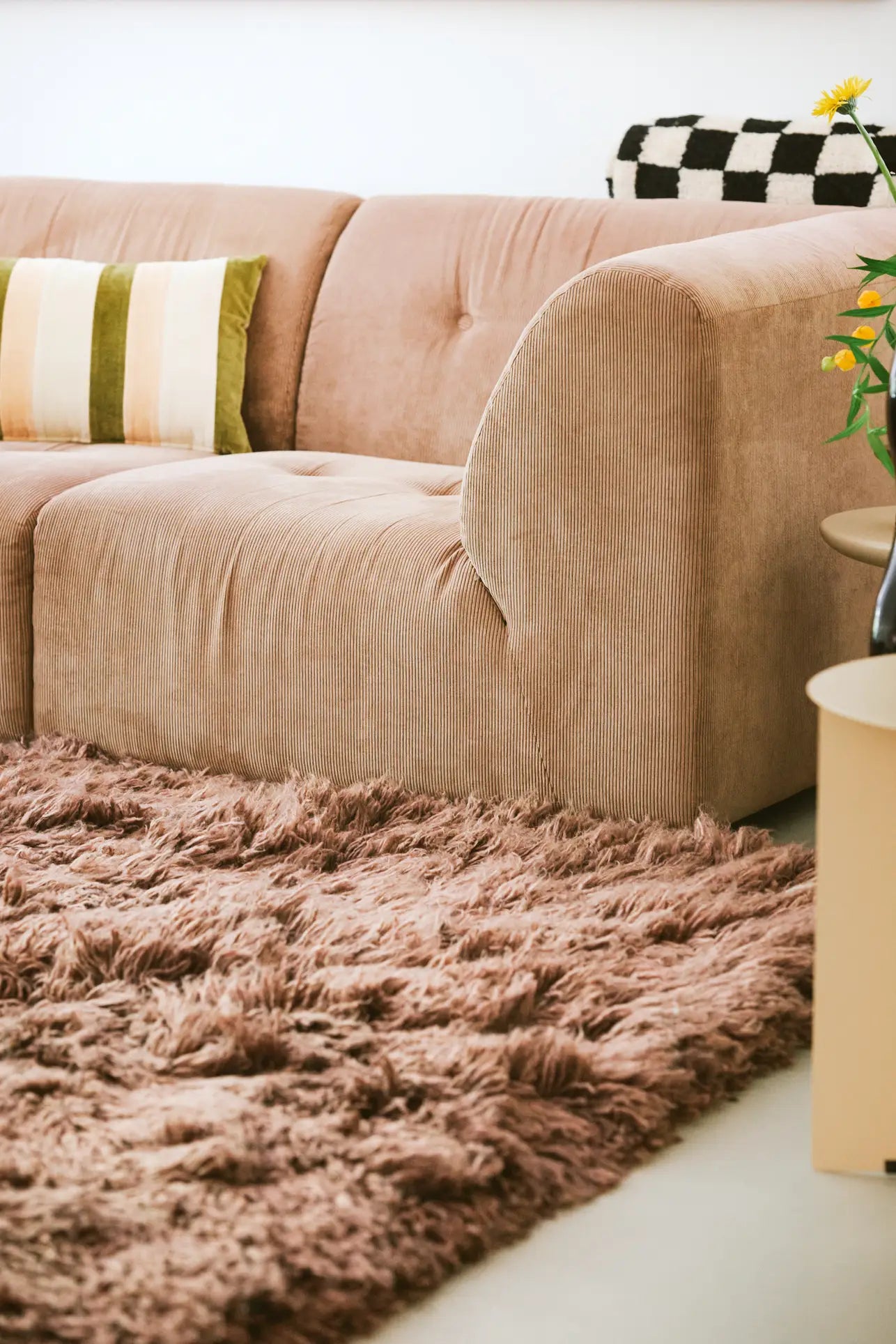 Create a cozy, customizable living space with the HKliving Vint Couch, Right by HKliving in light brown, styled with patterned cushions, a fluffy brown rug, a small round side table, and yellow flowers in a vase.