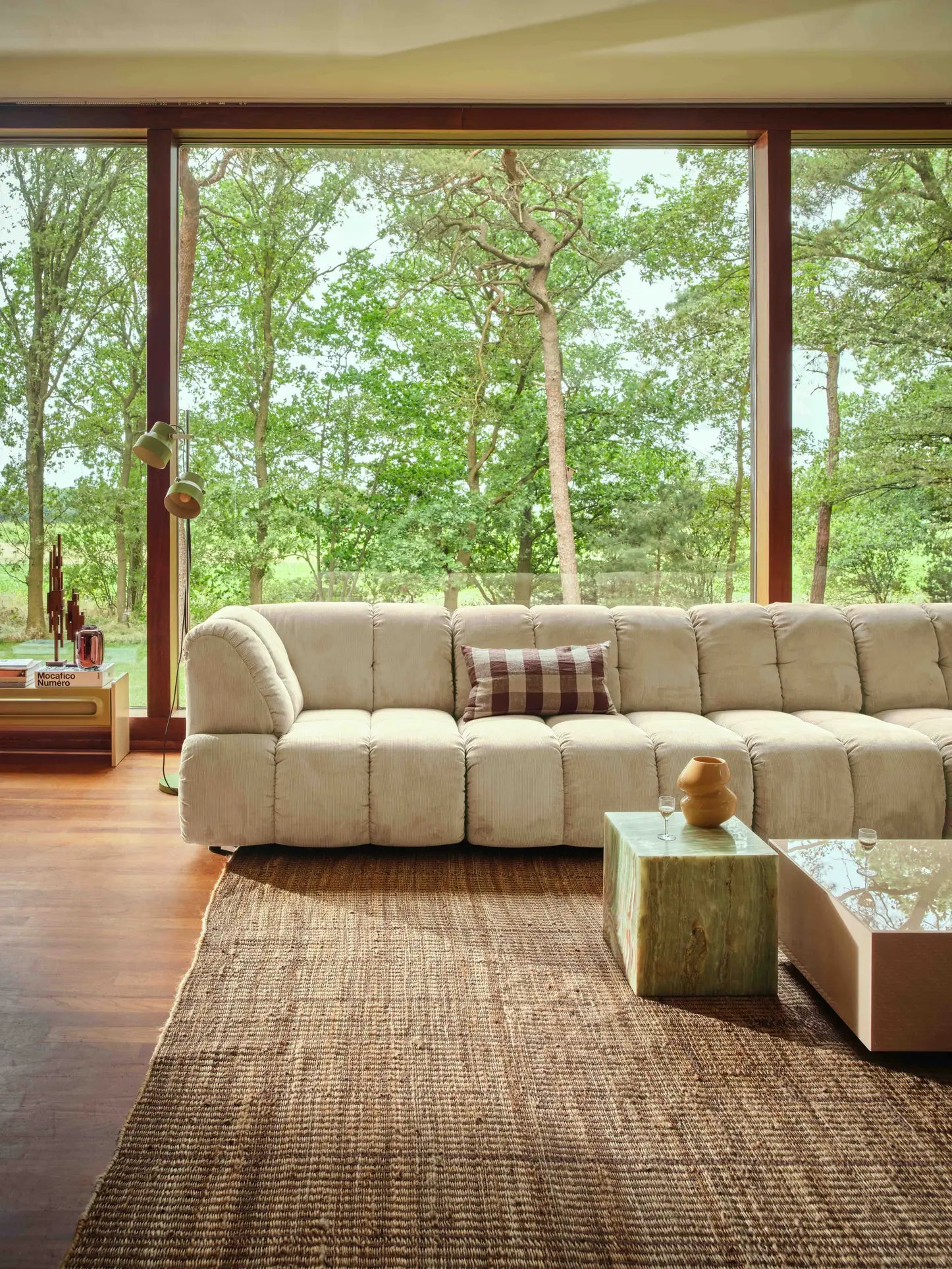 A cozy living room featuring the HKliving Wave Couch, Corner by HKliving with a plaid pillow, a wooden coffee table, a textured rug, and large floor-to-ceiling windows overlooking a lush green forest.