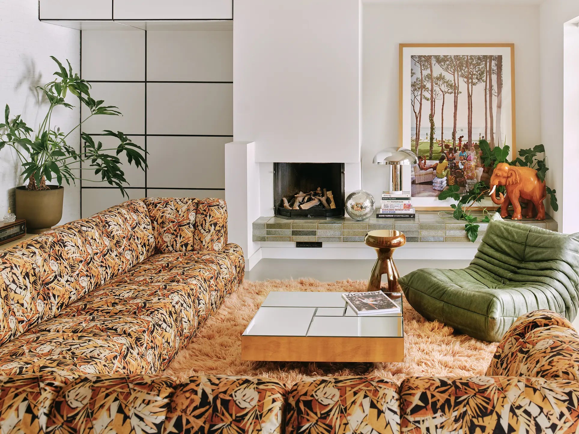 A retro-style living room features the HKliving Wave Couch, green lounge chair, shaggy orange rug, modern square coffee table, fireplace with plants, framed art, and a small orange mammoth figurine on the hearth.