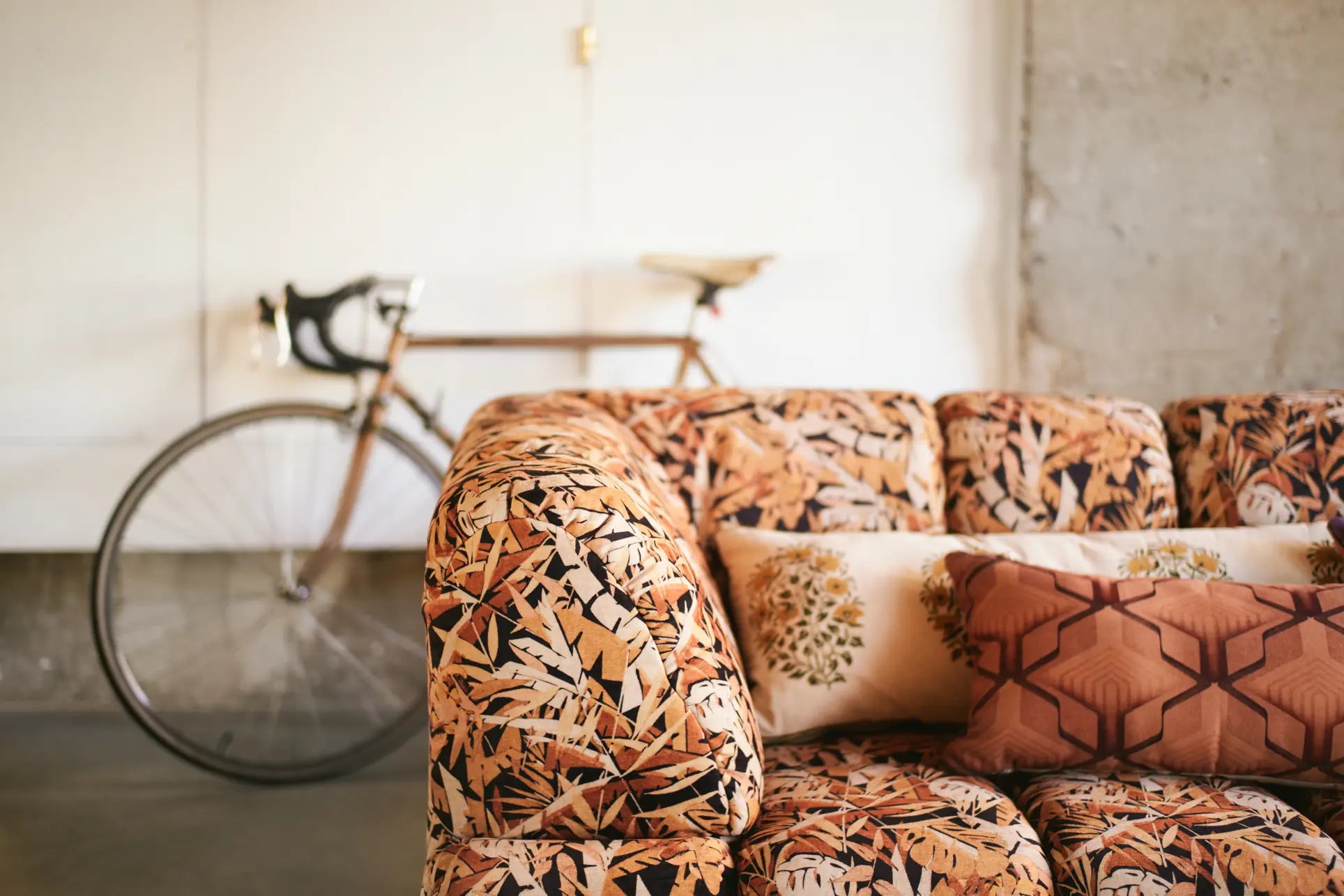 The HKliving Wave Couch, Corner by HKliving, with its patterned orange and brown fabric plus decorative pillows, stands out in the minimalist rooms foreground as a vintage bicycle rests against a white wall in the background.