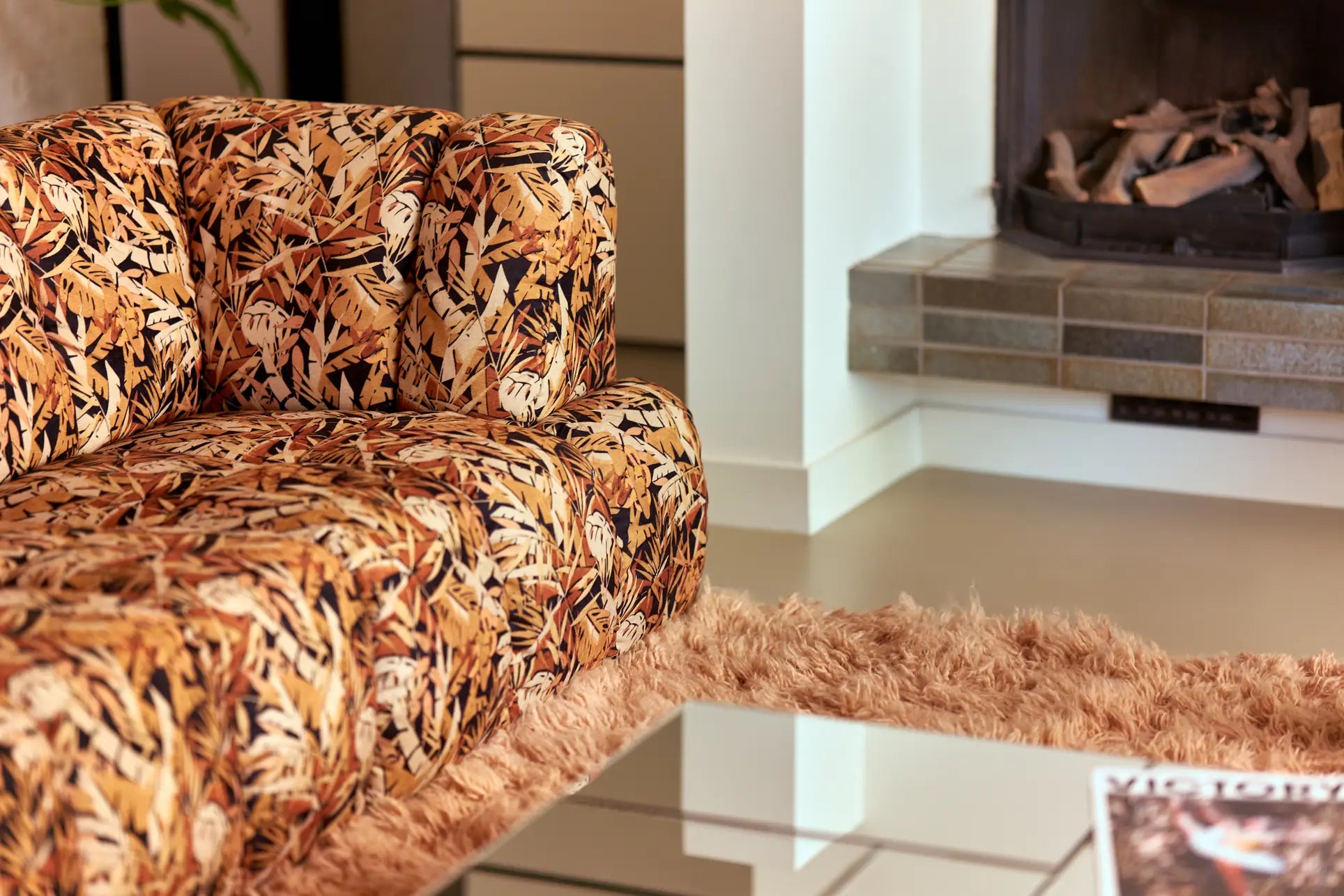 A close-up of the HKliving Wave Couch, Left High Arm by HKliving, featuring colorful autumn leaf-patterned fabric. It sits on a shaggy beige rug beside a glass coffee table, with a fireplace visible in the background.