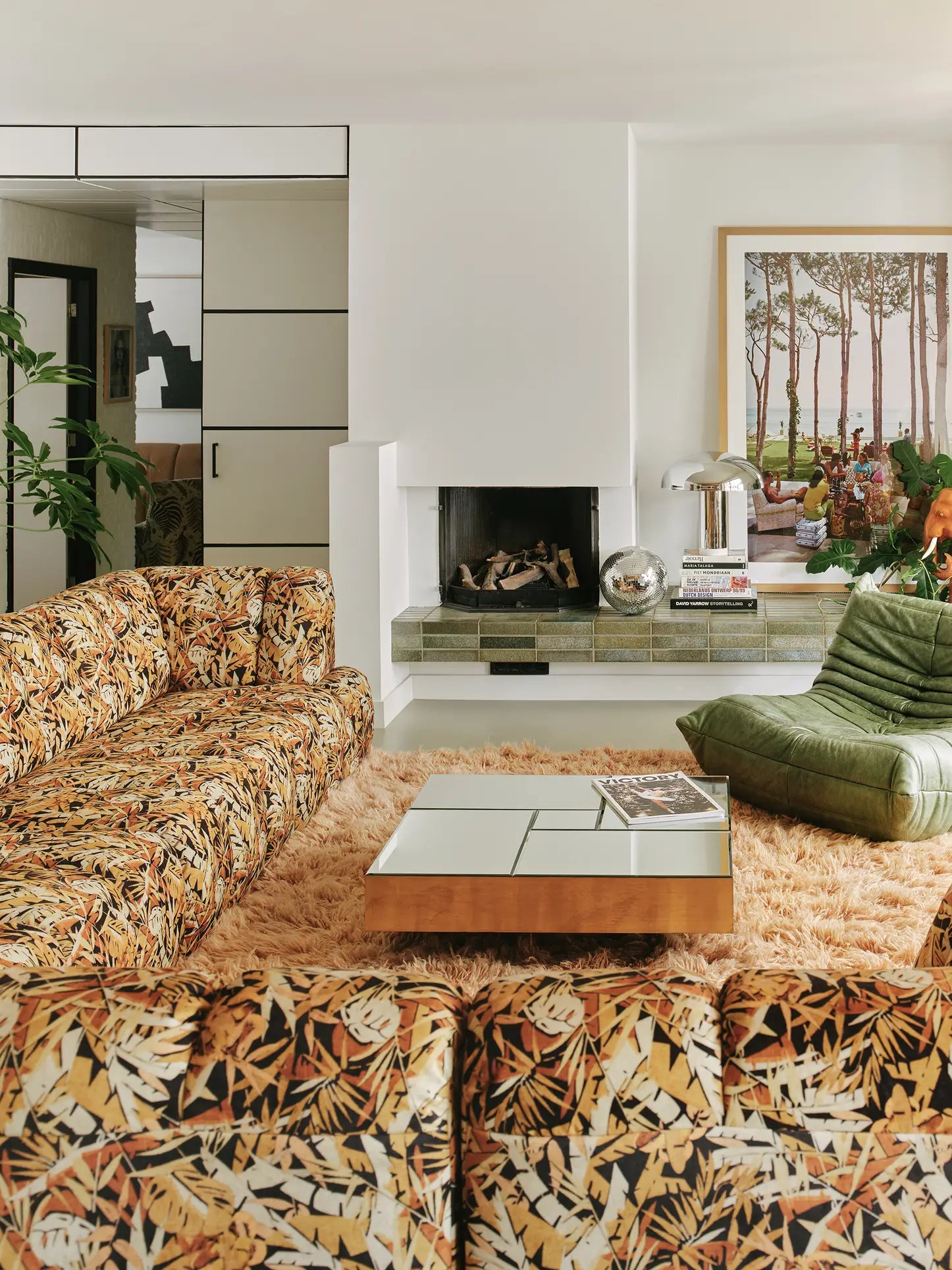 A retro living room with patterned sofas, a green lounge chair, an HKliving Wave Couch (Left High Arm), a square coffee table, shaggy orange rug, fireplace, and framed landscape photo. Modern decor accents the bright, cozy space.
