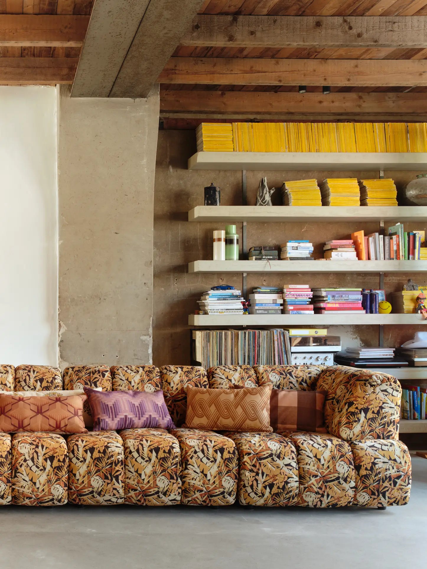 The HKliving Wave Couch, Left High Arm by HKliving with assorted pillows sits before shelves filled with yellow magazines, books, records, and decor in a room featuring exposed wooden beams and concrete accents.