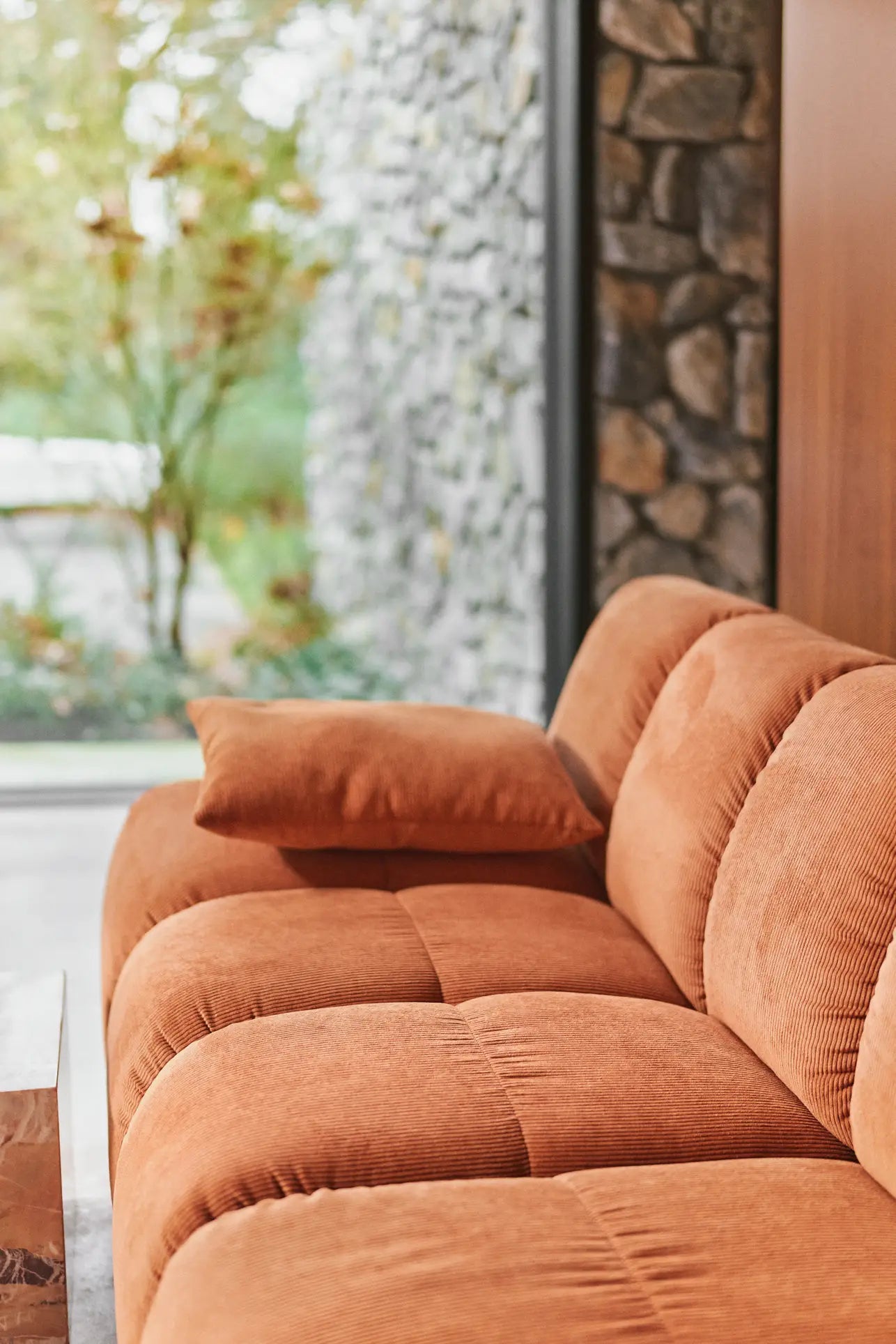 A close-up of the HKliving Wave Couch, Left Low Arm in orange corduroy with a matching cushion, set in a stylish living room beside a stone wall and large window—perfect for customizing your space with HKliving style.