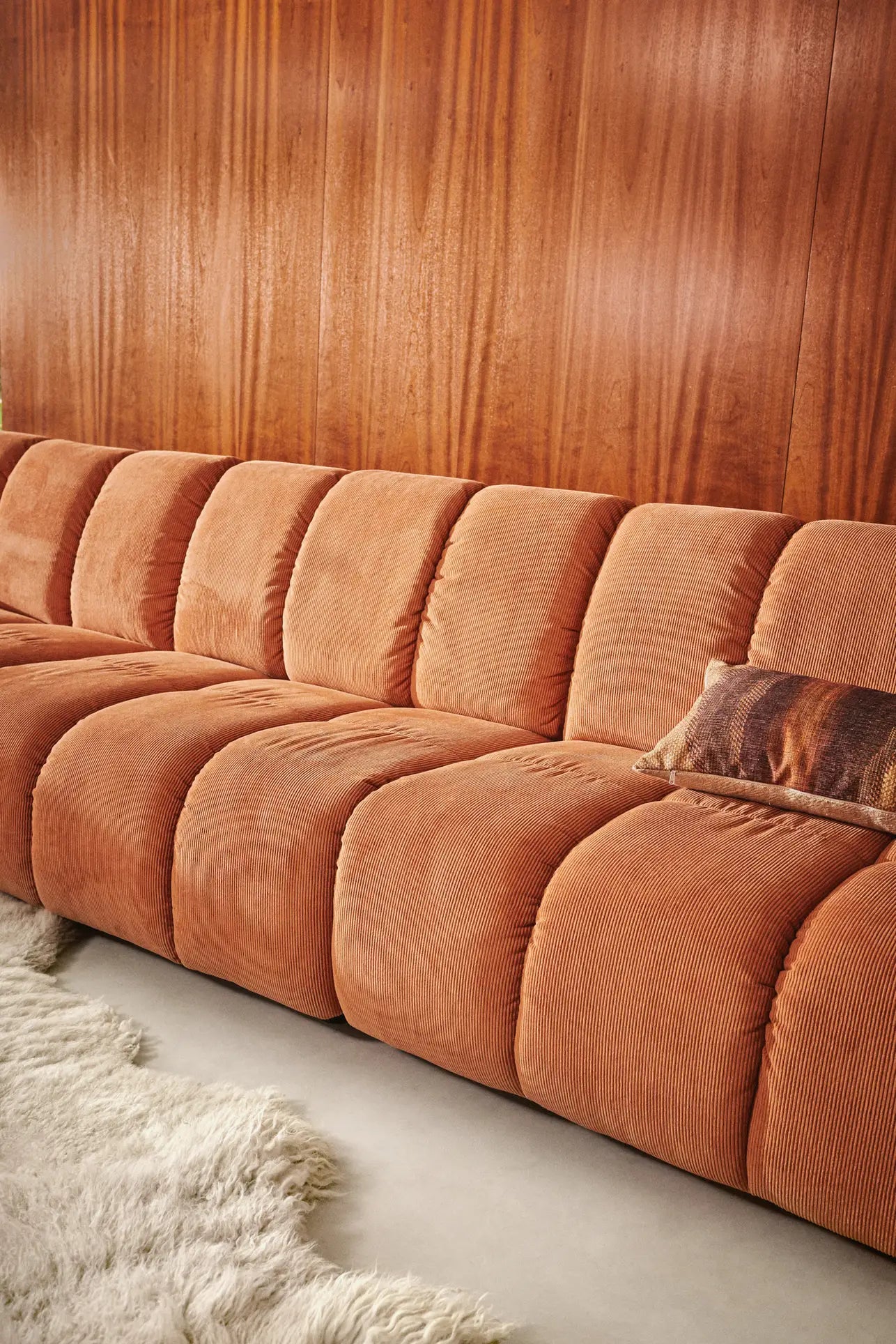 The HKliving Wave Couch, Middle by HKliving features a rust hue and modern design, set against wood paneling with a fuzzy cream rug and decorative pillow, creating a cozy 1970s-inspired atmosphere.