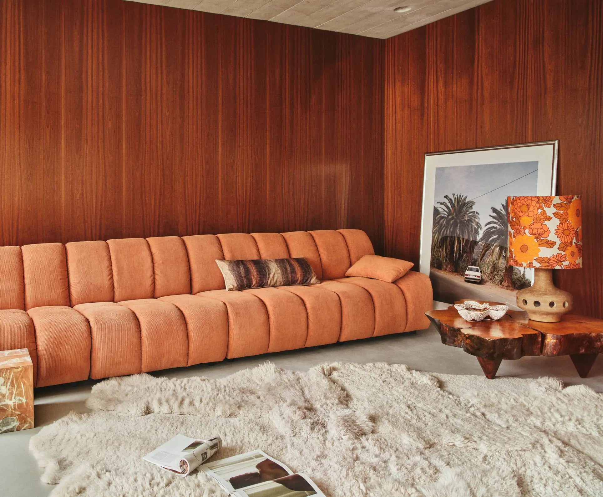 A cozy living room with a peach HKliving Wave Couch, a furry white rug, wooden coffee table, retro orange lamp, and framed palm tree photo against dark wood-paneled walls exudes 1970s design.