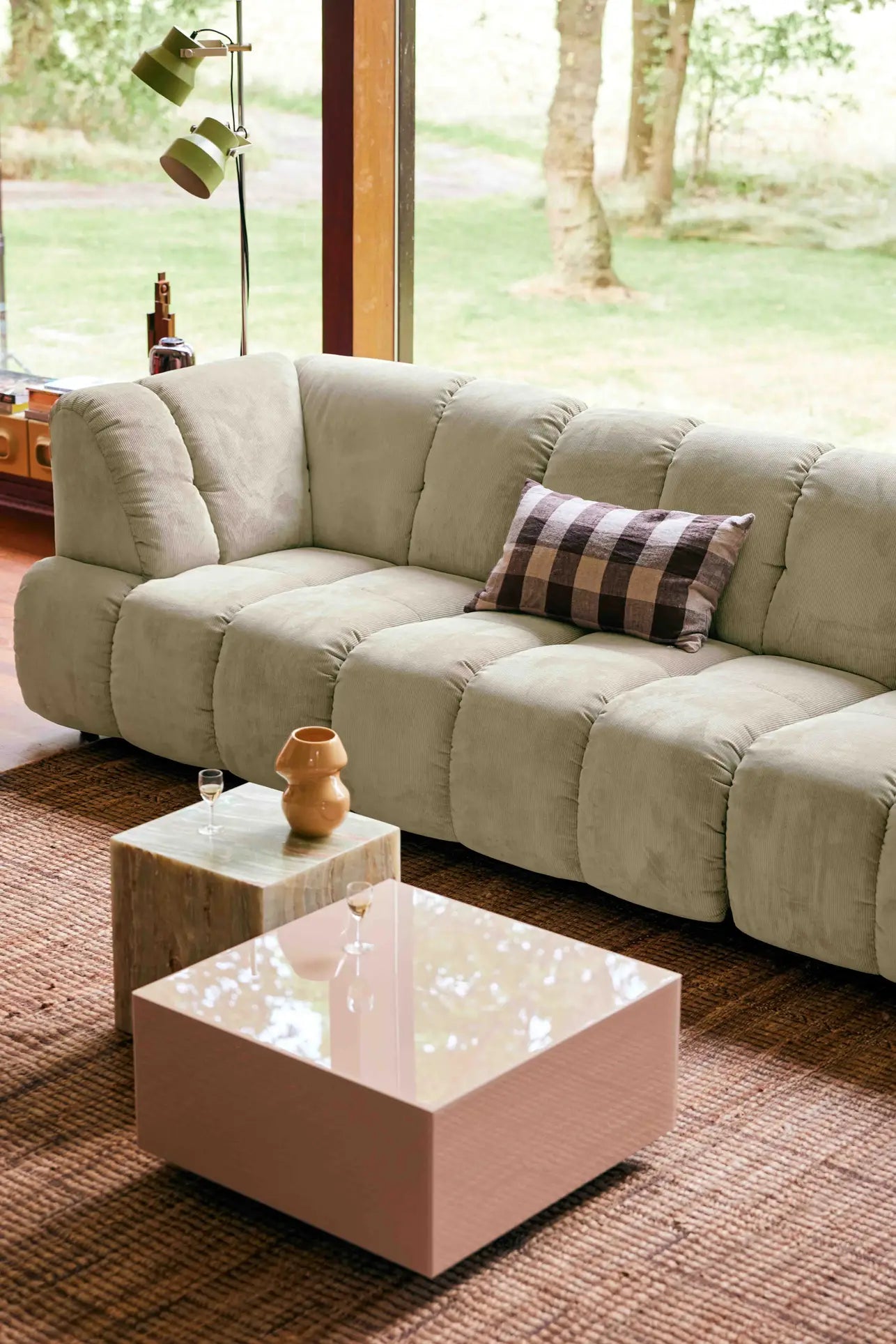 The HKliving Wave Couch, Middle in beige with a plaid pillow is set by large windows with yard views. A woven rug, two sleek coffee tables, and a ceramic vase complete the inviting living room by HKliving.