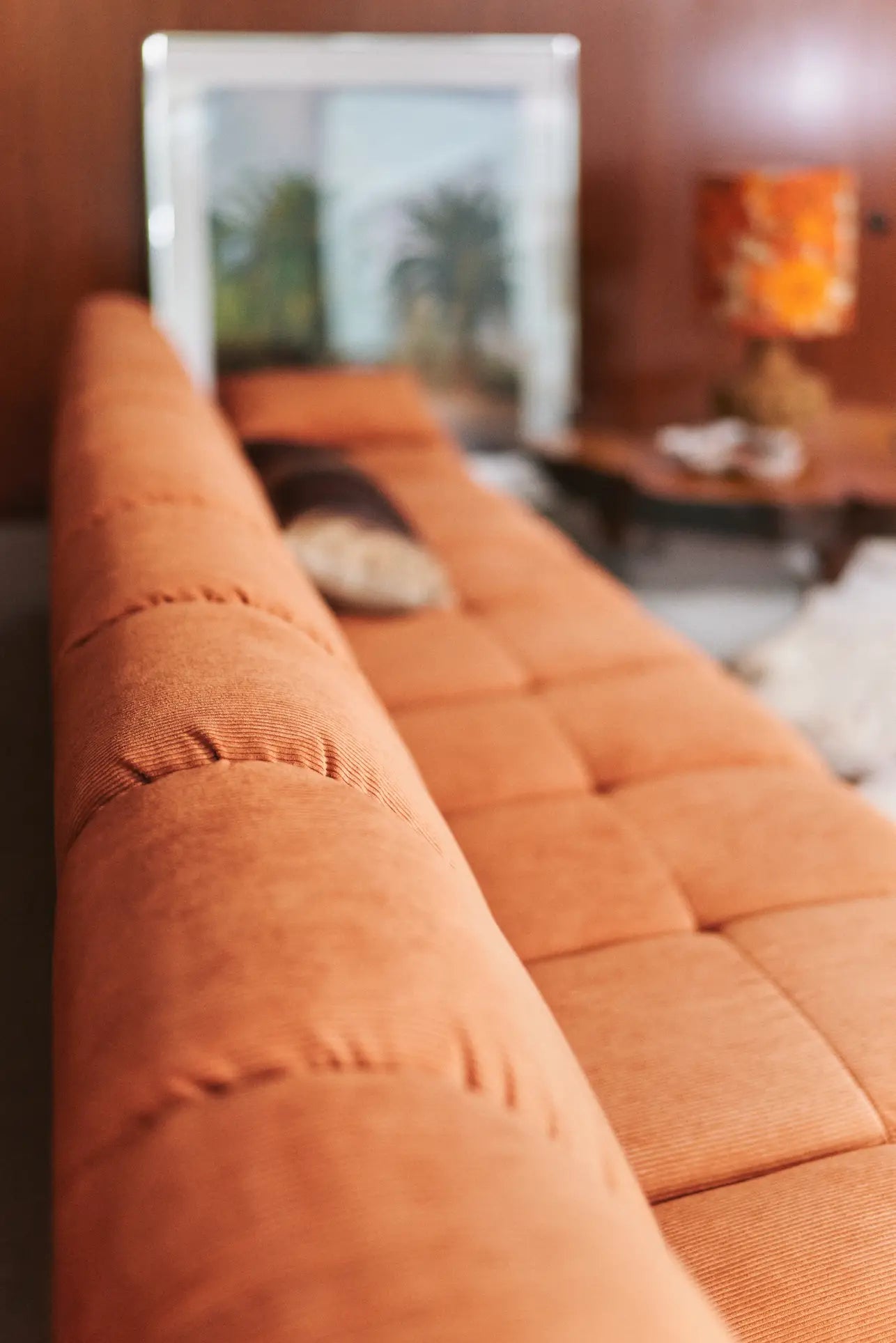 A close-up of the HKliving Wave Couch, Middle Small by HKliving in orange tufted fabric, set in a retro living room with a blurred 1970s-style wood-paneled wall, table lamp, and framed photo in the background.
