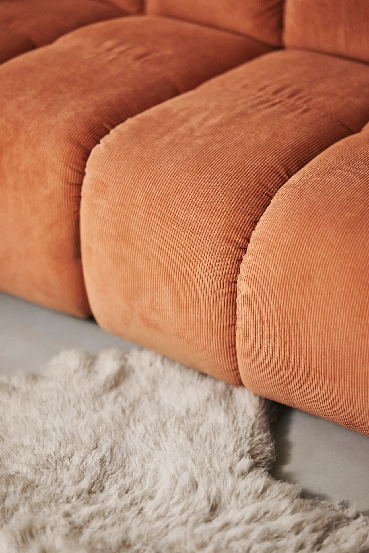 Close-up of the HKliving Wave Couch, Middle Small by HKliving in orange corduroy with soft, rounded cushions next to a white fluffy rug on a light-colored floor.