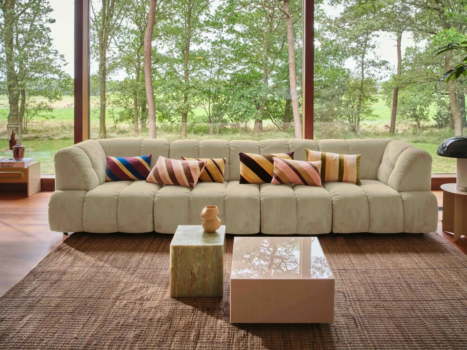 The HKliving Wave Couch, Middle Small by HKliving, in beige with colorful striped pillows, sits on a woven rug before floor-to-ceiling windows with a view of green woods; two modern coffee tables are in front of this customizable sofa.