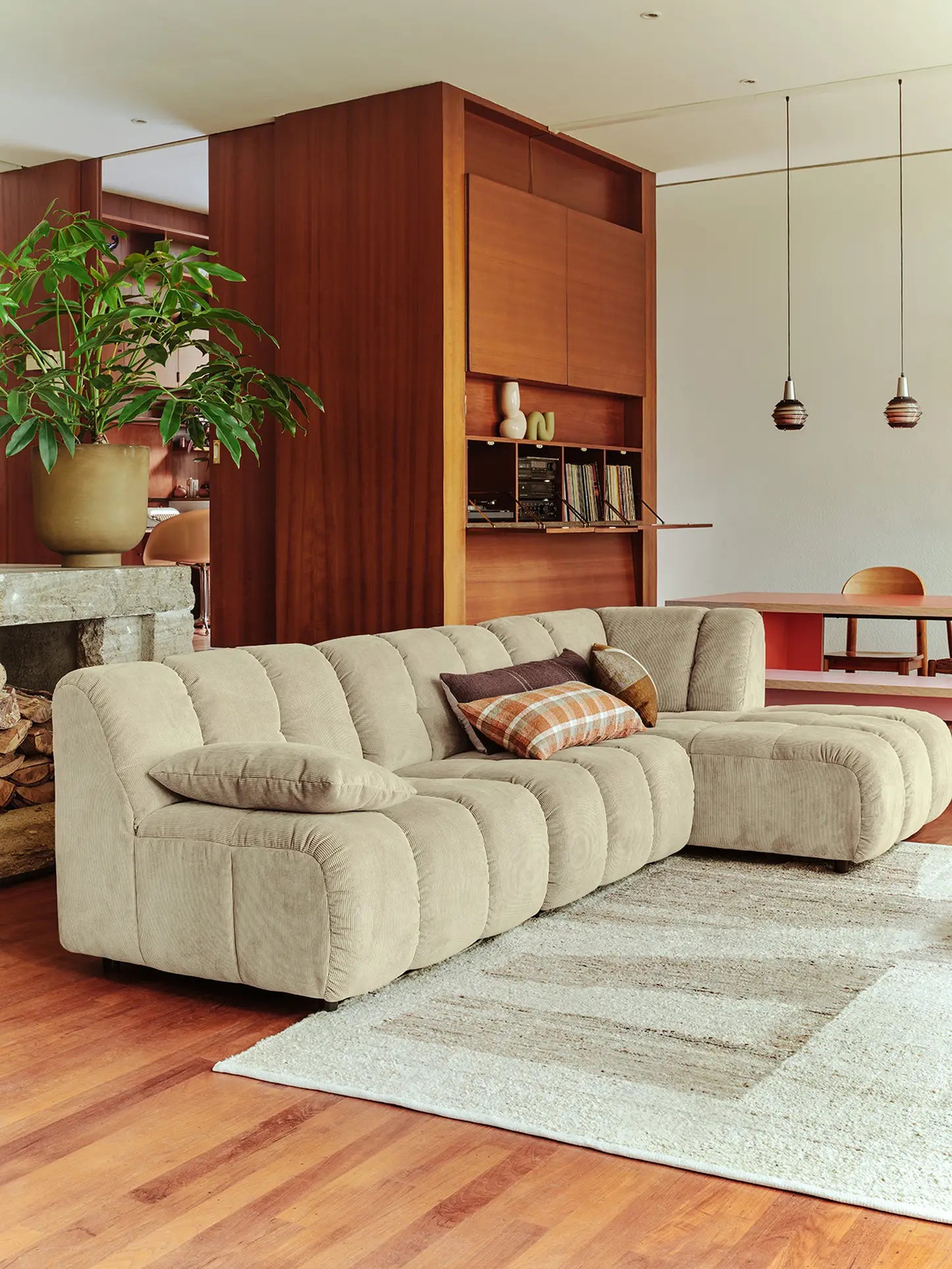 A modern living room showcases the HKliving Wave Couch, Right Low Arm by HKliving with patterned pillows, a light rug on wood floors, a potted plant, built-in shelving, and pendant lights over a red dining table in the background.