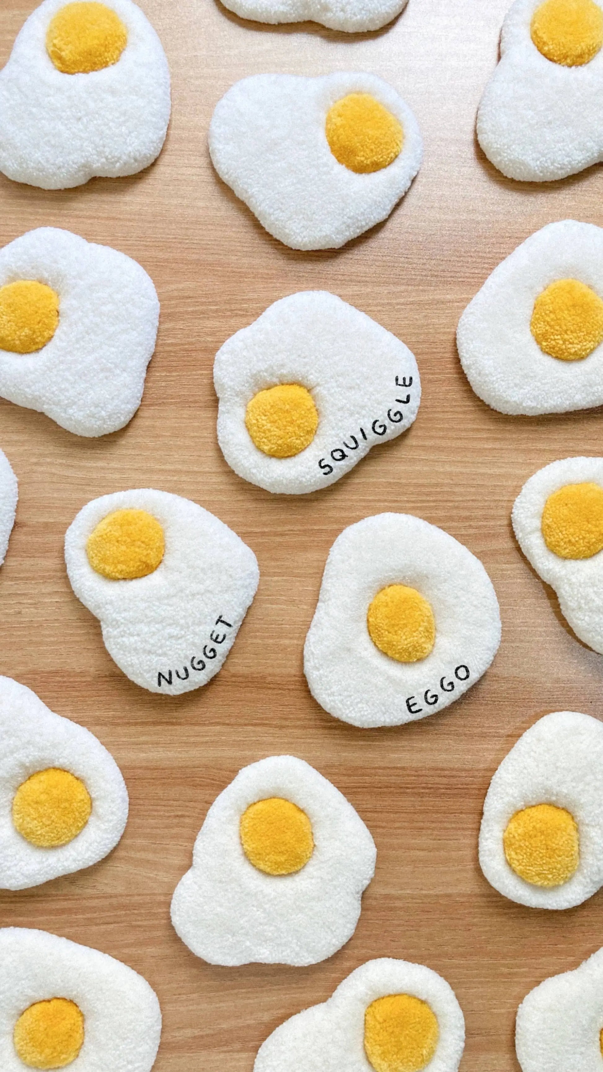 Habichl Tufted Egg Coasters by Habichl, shaped like sunny-side-up eggs, are displayed on wood. Names like NUGGET, SQUIGGLE, and EGGO appear on the whites, giving these playful coasters a handcrafted, plush toy look.