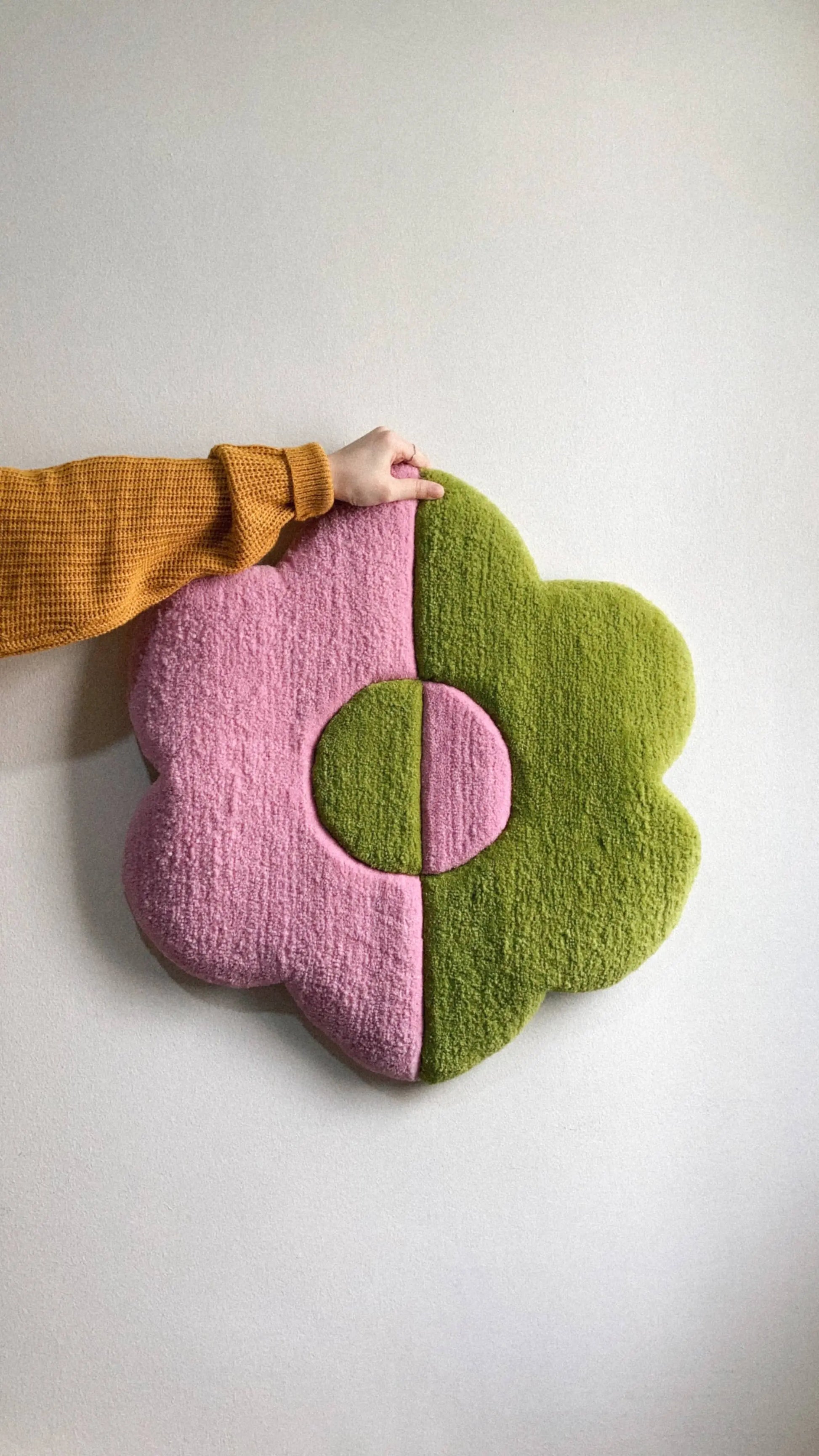 Someone in a mustard yellow sweater touches a Habichl Tufted Flower Floor Rug by Habichl, displayed on the wall. This playful mat features pink and green halves with a coordinating multicolored center circle.