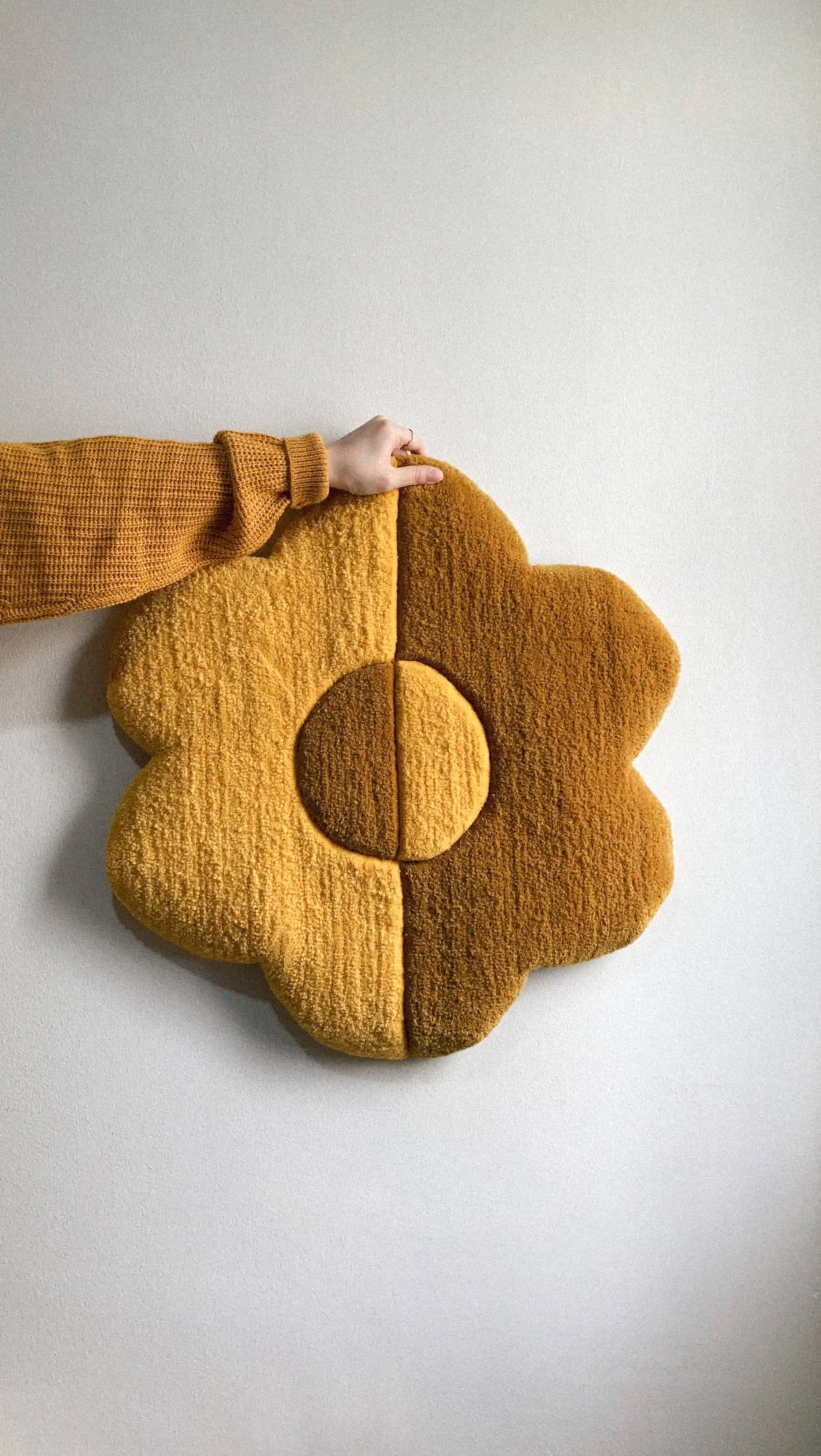 A hand in a brown sweater holds the Habichl Tufted Flower Floor Rug, a plush, flower-shaped piece by Habichl with yellow and brown halves, set against a plain light-colored wall.