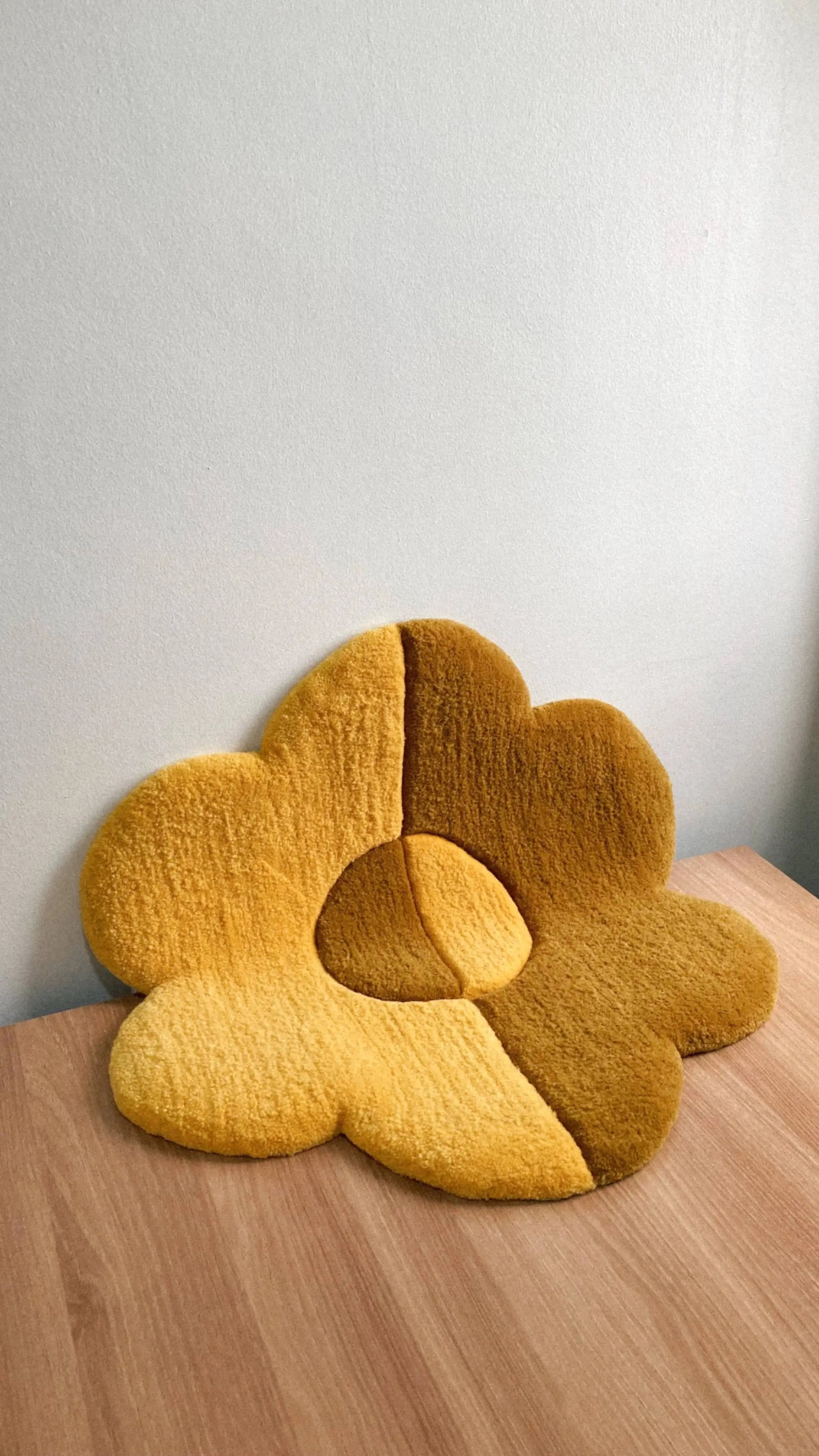The Habichl Tufted Flower Floor Rug, a plush yellow-and-brown cushion with petal-like sections, adds a whimsical touch as it rests on a light wooden table against a plain pale wall.