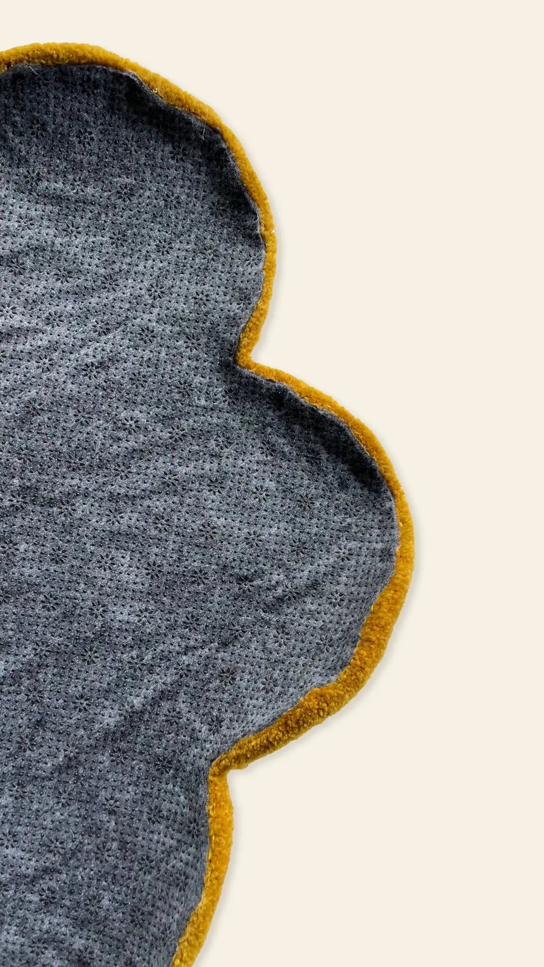 A close-up of a gray, flower-shaped fabric with a yellow border, set on a beige background. Only part of the whimsical Habichl Tufted Flower Floor Rug by Habichl is visible in the image.