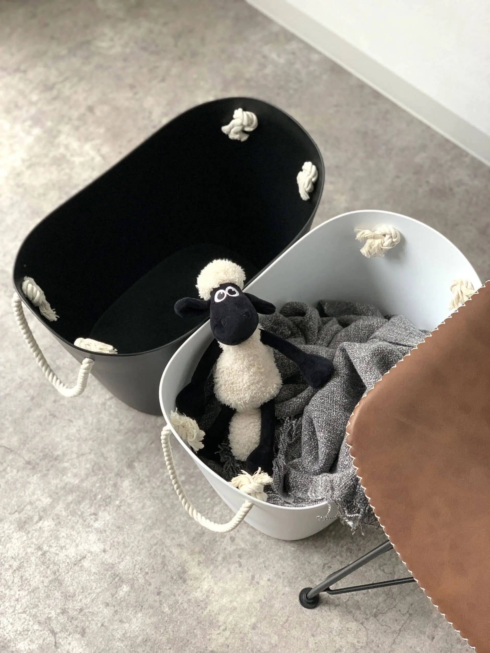 A black bin and a white Hachiman Balcolore Storage Basket M with rope handles sit on the floor. The minimalist Japanese-inspired basket holds a gray blanket and plush sheep toy next to a brown leather chair.